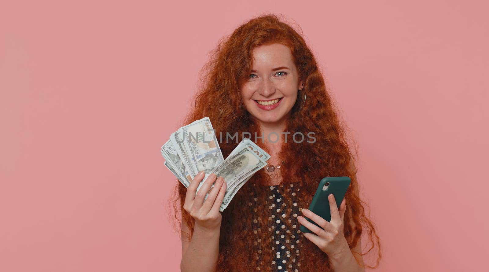 Happy redhead young woman looking smartphone display sincerely rejoicing win, receiving money dollar cash banknotes, success lottery luck. Ginger girl indoors studio isolated on pink wall background