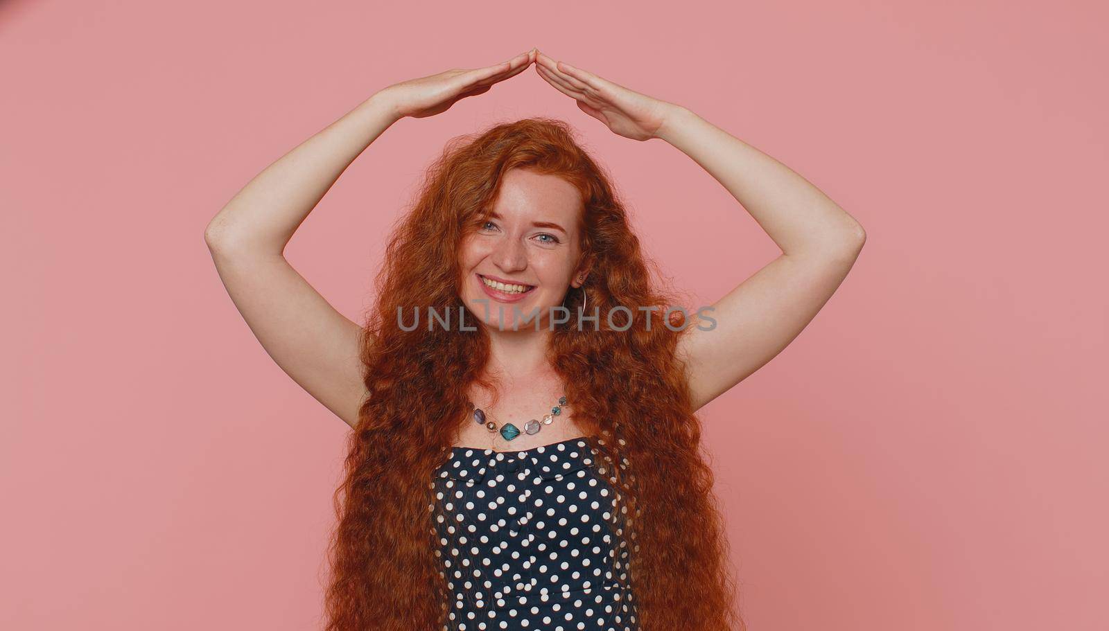 Young redhead woman playing childish catching up game, feeling in safe making roof above head with hands, insurance, security service. Ginger girl with freckles isolated on pink studio background