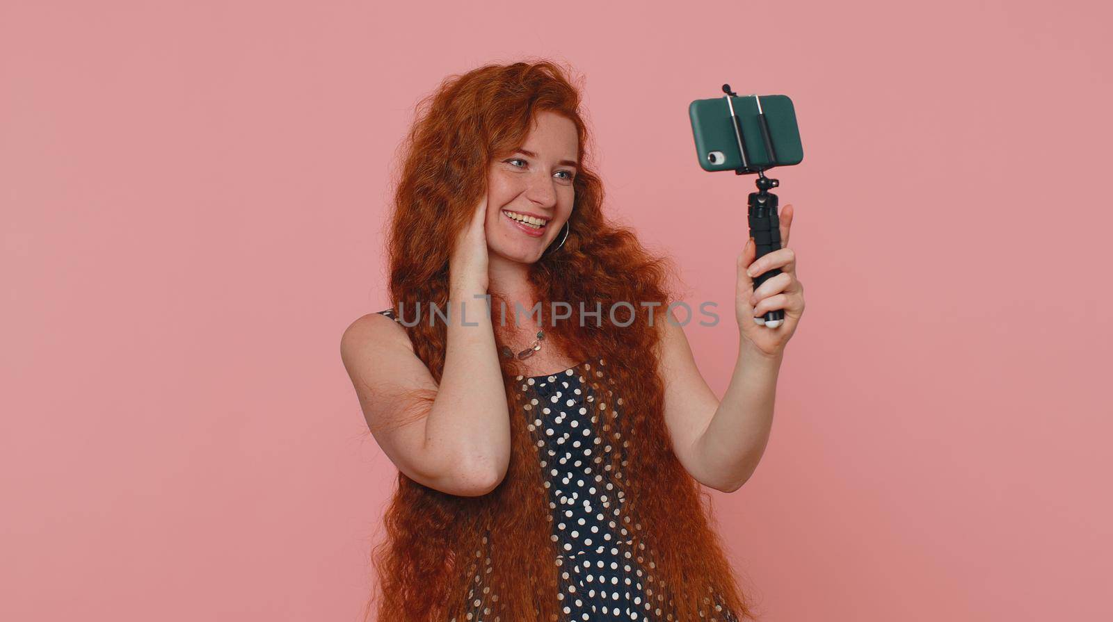 Redhead woman in dress traveler blogger taking selfie on mobile phone selfie stick, communicating video call online with subscribers. Young ginger girl isolated alone on pink studio background