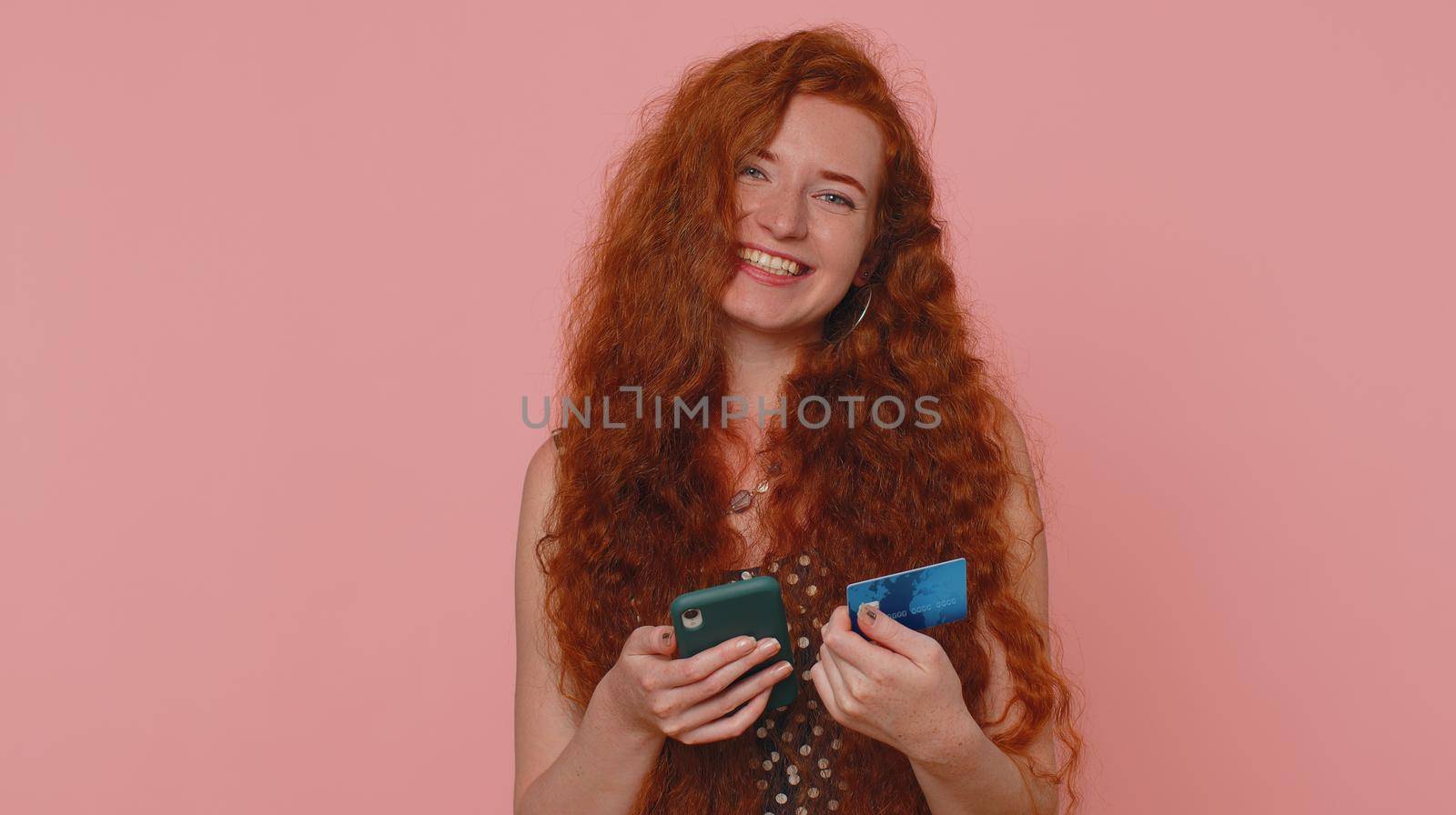 Sincere redhead woman customer using credit bank card and smartphone while transferring money, purchases online shopping, payment. Internet finance. Young girl isolated alone on pink wall background