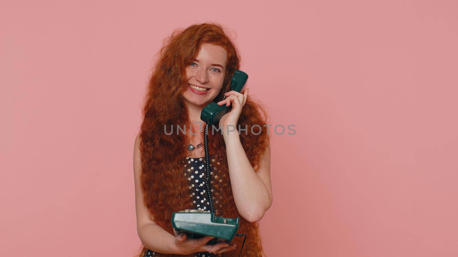 Hey You Call Me Back. Cheerful ginger woman secretary in dress talking on wired vintage telephone of 80s, says hey you call me back. Young redhead freckles face girl isolated on pink studio background