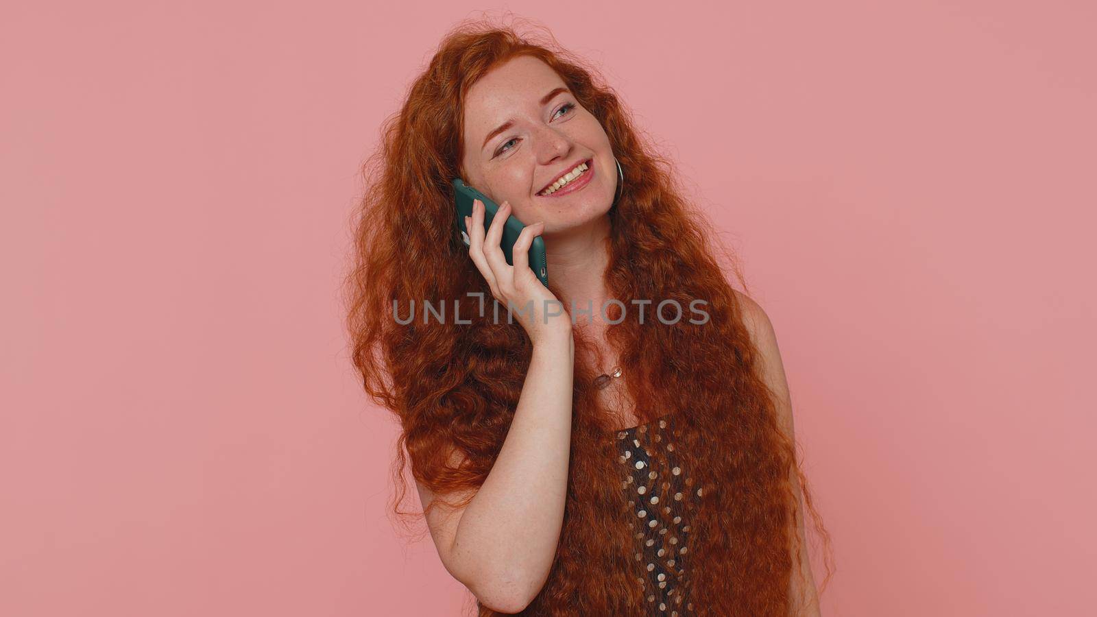 Redhead young woman in polkadot dress having pleasant mobile conversation using smartphone with friends, telling good news. Adult ginger freckles face girl indoors isolated on pink studio background