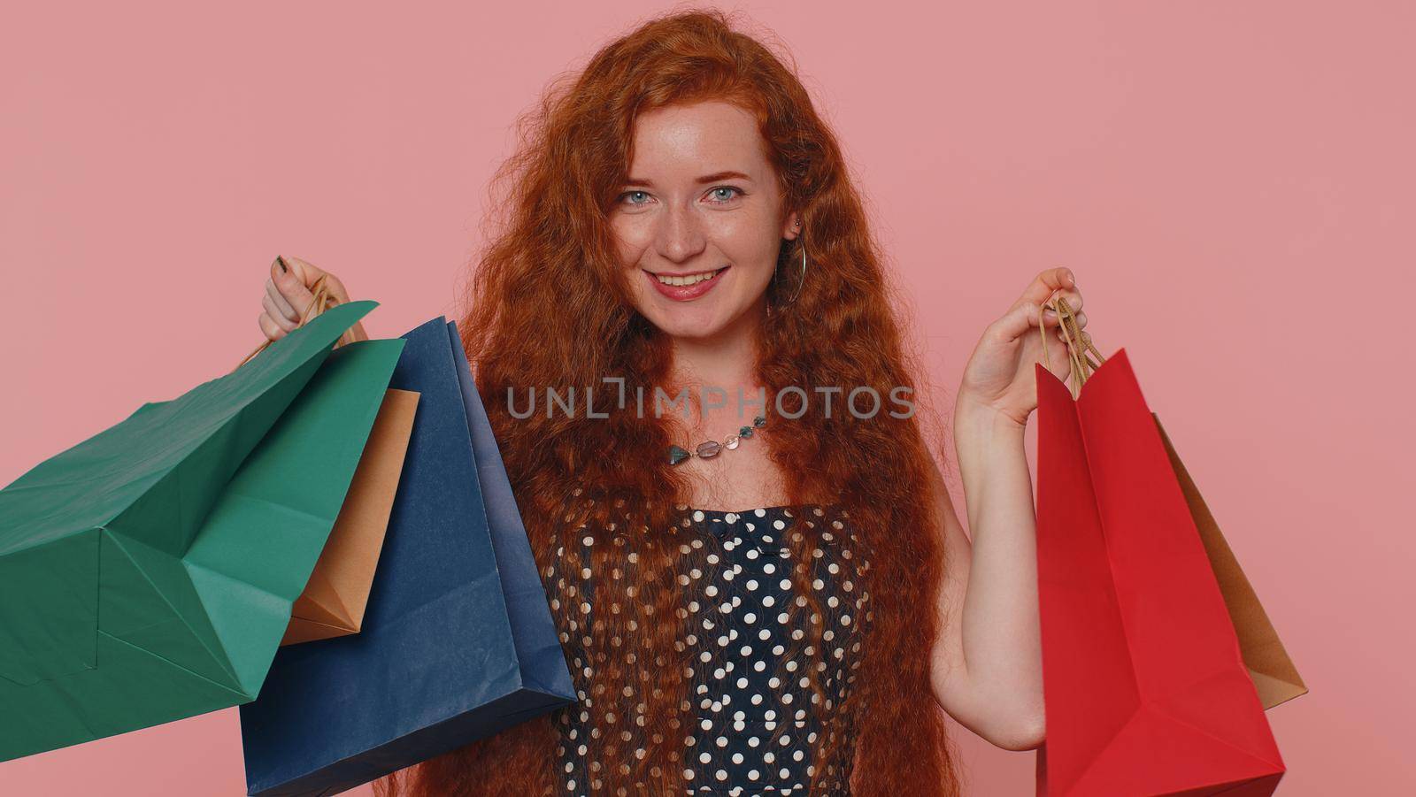 Happy redhead shopaholic woman showing shopping bags, advertising discounts, smiling looking amazed with low prices shopping on Black Friday holidays. Ginger girl indoors isolated on pink background