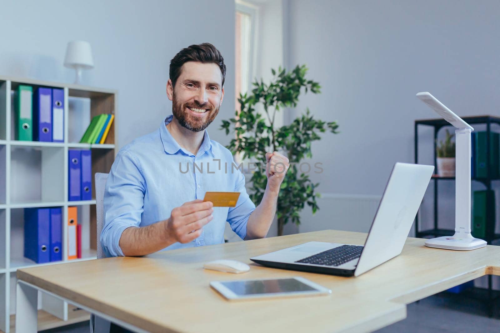 A young man at home looks at the camera and smiles holding a credit bank card for online shopping in the store, rejoices a successful buyer uses a laptop