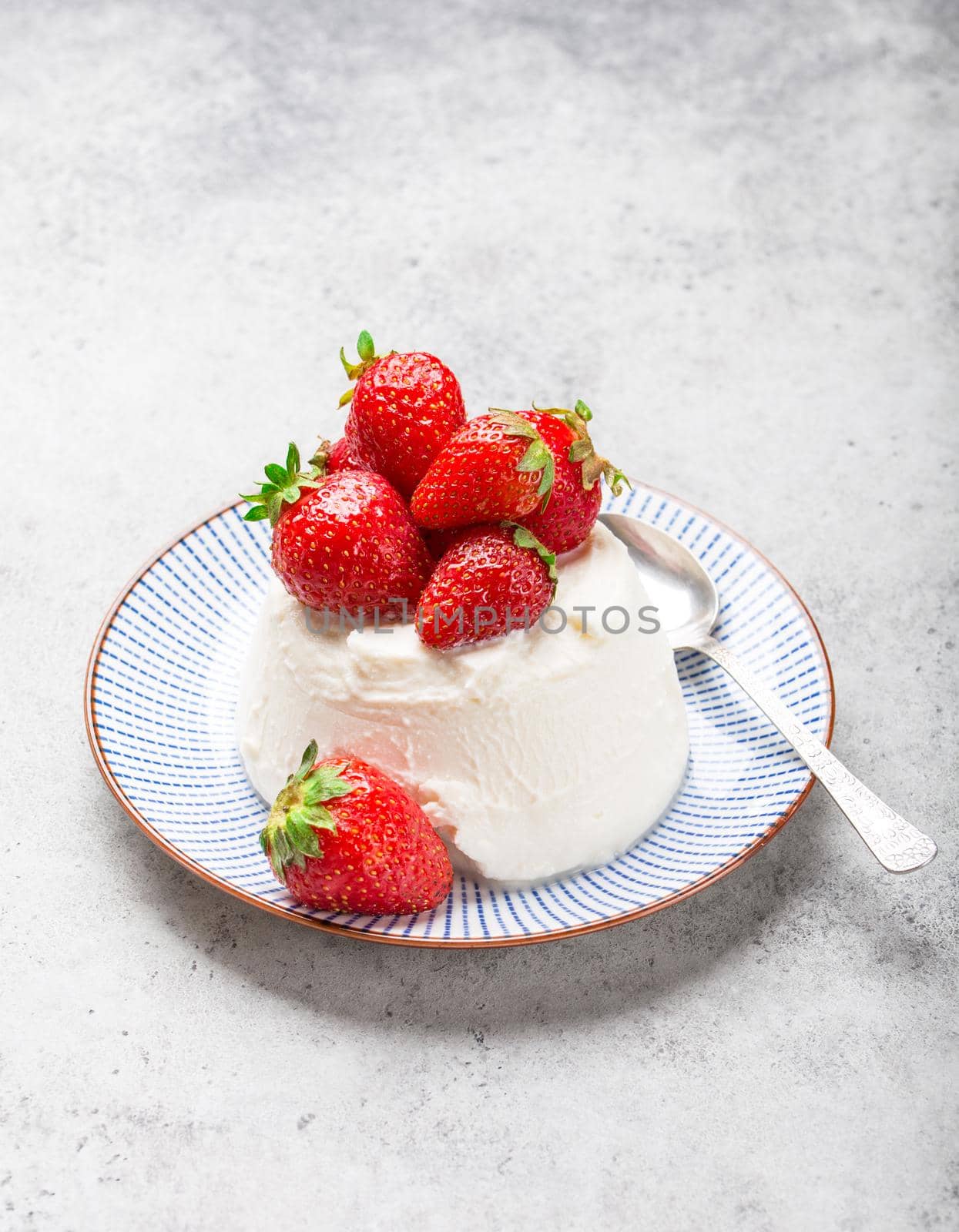 Fresh Italian cheese ricotta with strawberries on a plate with a spoon, grey rustic stone background, light summer dessert or snack, good for diet or healthy eating. Selective focus, close-up