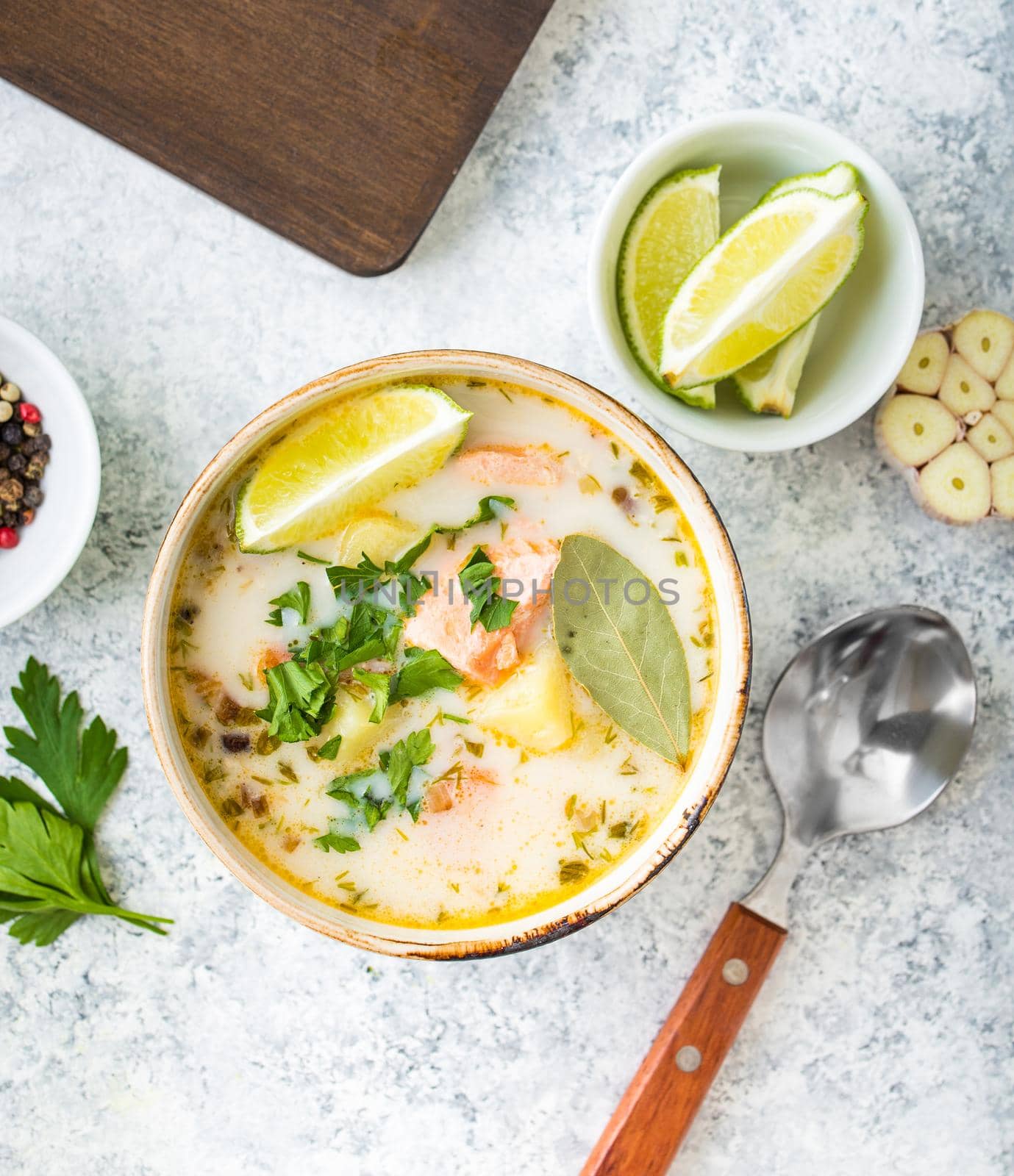 Salmon soup with potatoes, carrot, cream, herbs. Scandinavian/Norwegian fish soup in bowl, cutting wooden board, spoon, rustic white concrete background. Salmon soup for dinner. Top view. Close-up