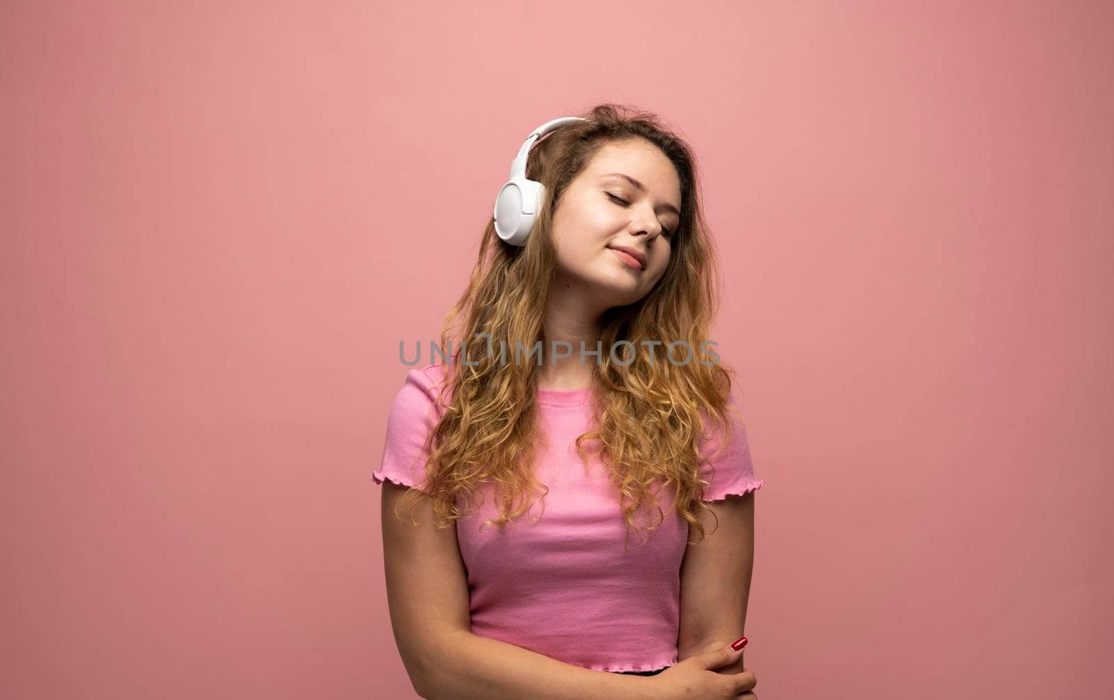 Portrait of charming curly woman in white massive headphones listening to music on isolated pink background. Curly girl in t-shirt smiles. by vovsht