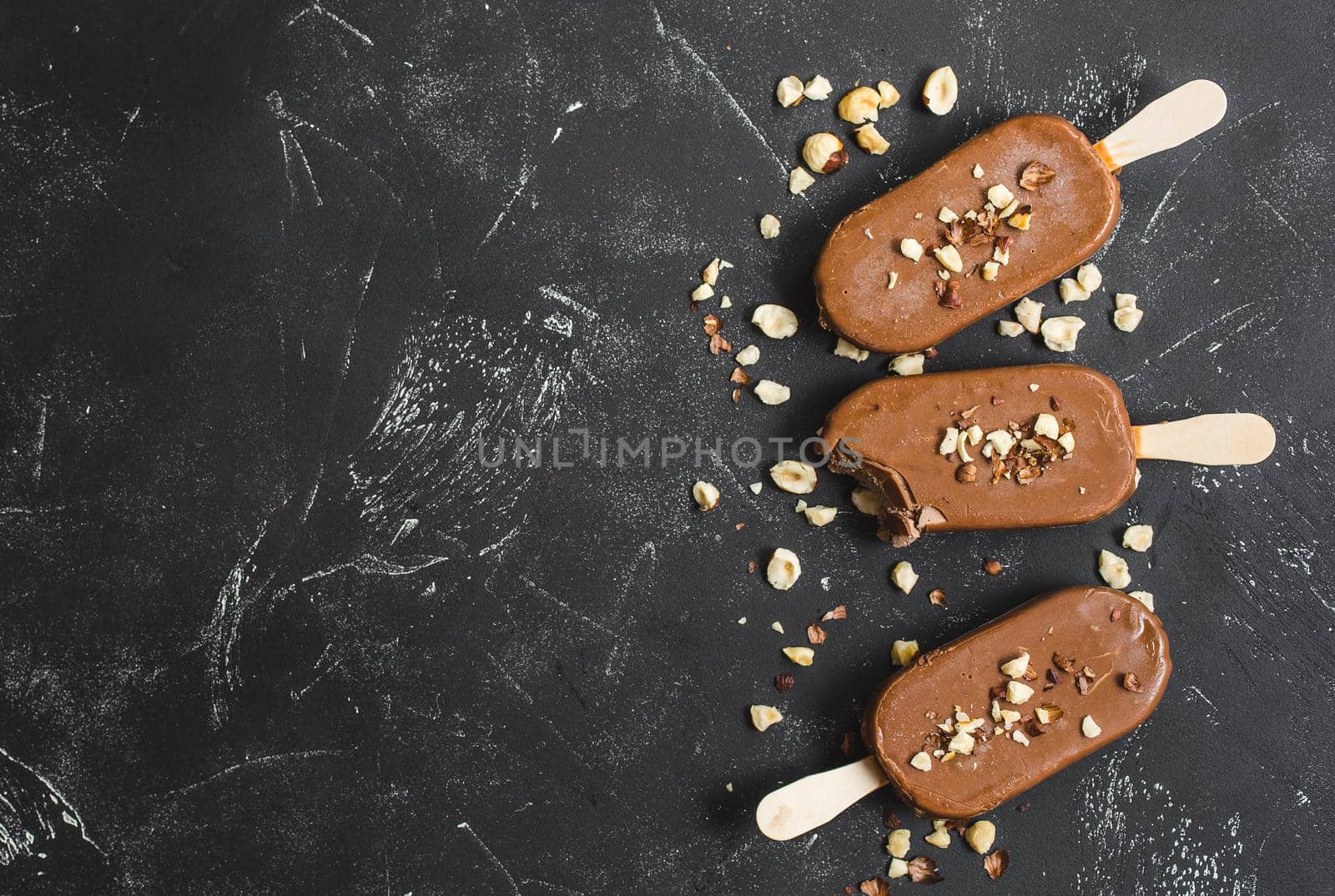 Milk chocolate popsicles with hazelnuts. Close-up. Ice cream popsicles covered with chocolate, sticks, black stone background. Space for text. Top view. Chocolate ice cream bars, nuts. Bitten popsicle