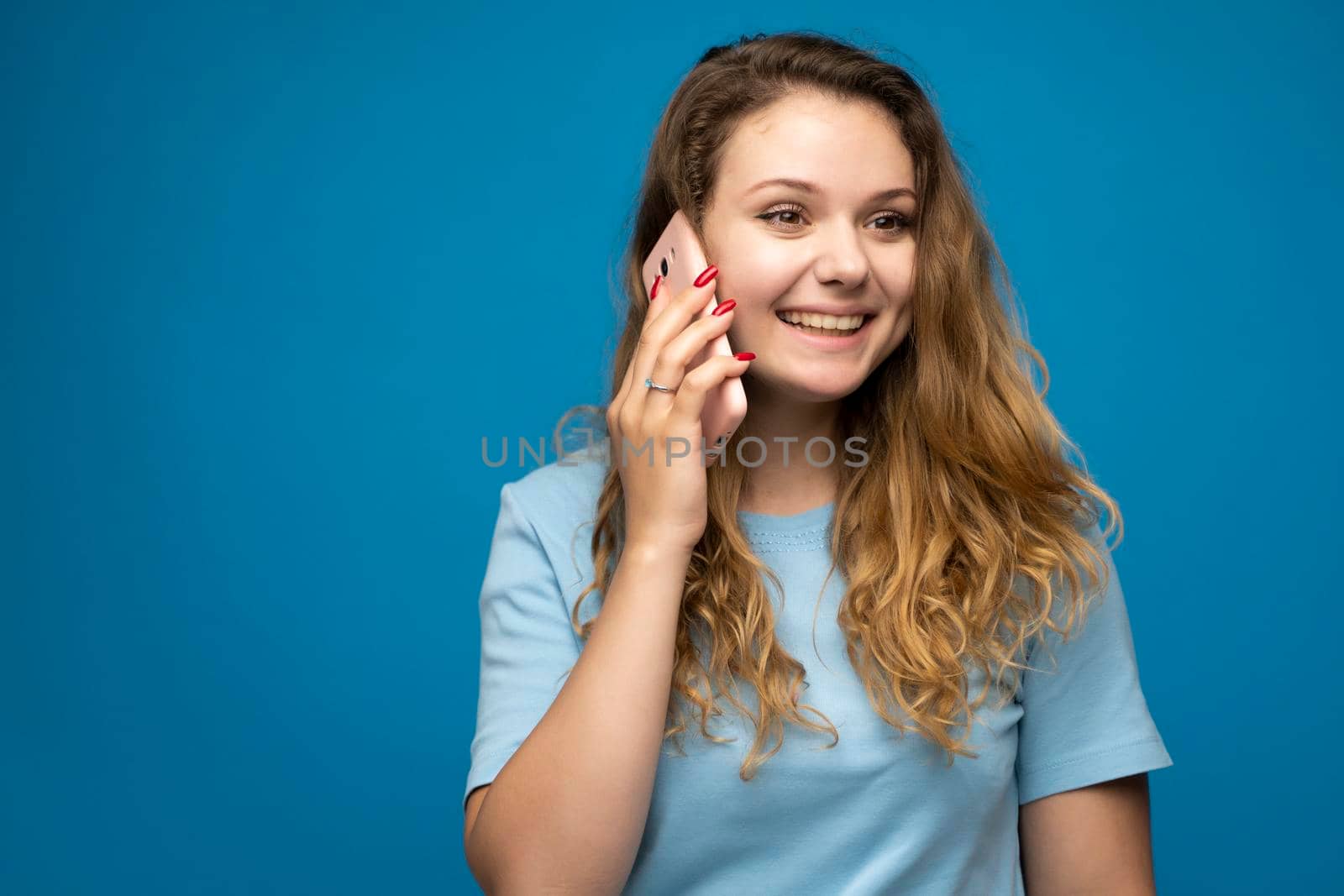 Young smiling happy fun woman in blue basic casual t-shirt talking speak on mobile phone conducting pleasant conversation on blue background studio portrait