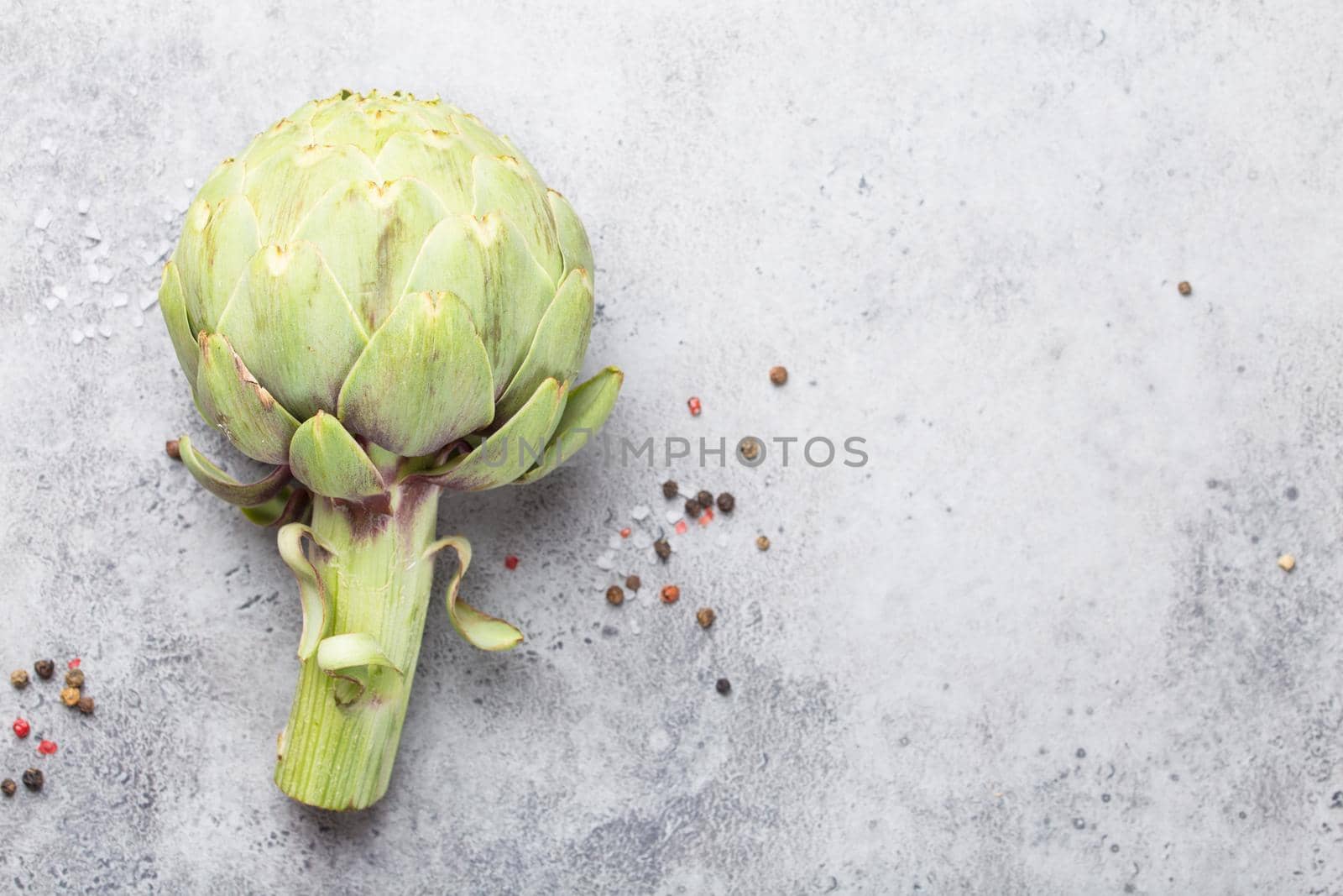 Close-up of raw fresh artichoke with seasonings ready to cook, great as vegetarian food or ingredient for healthy salads and diets, grey stone rustic background, top view. Space for text