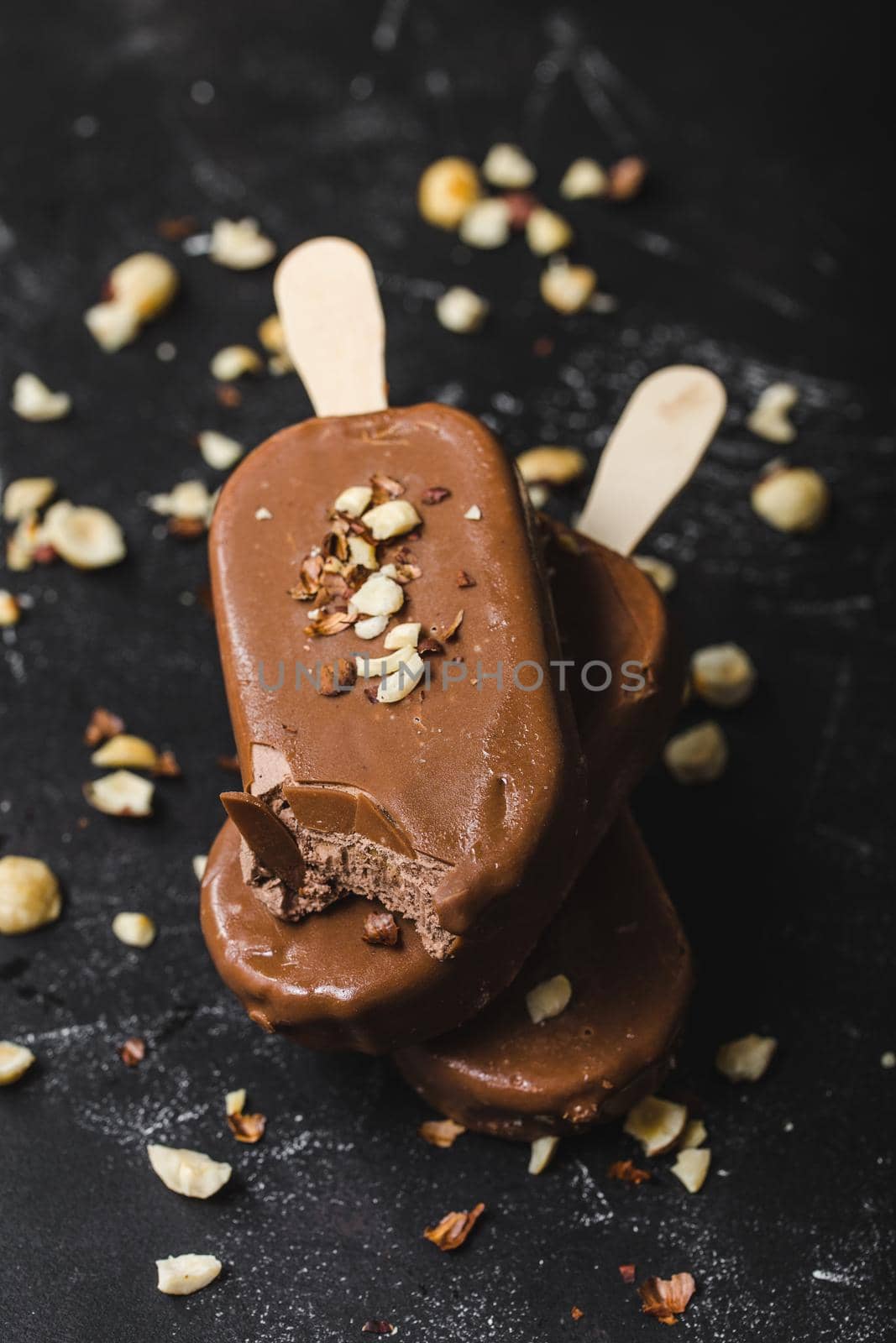 Milk chocolate popsicles with hazelnuts. Close-up. Ice cream popsicles covered with chocolate, sticks, black stone background. Selective focus. Chocolate ice cream bars, nuts. Dessert. Bitten popsicle