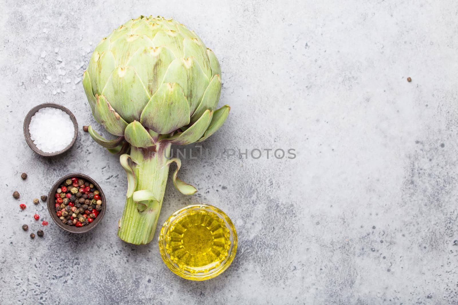 Close-up of raw fresh artichoke with seasonings ready to cook, great as vegetarian food or ingredient for healthy salads and diets, grey stone rustic background, top view. Space for text