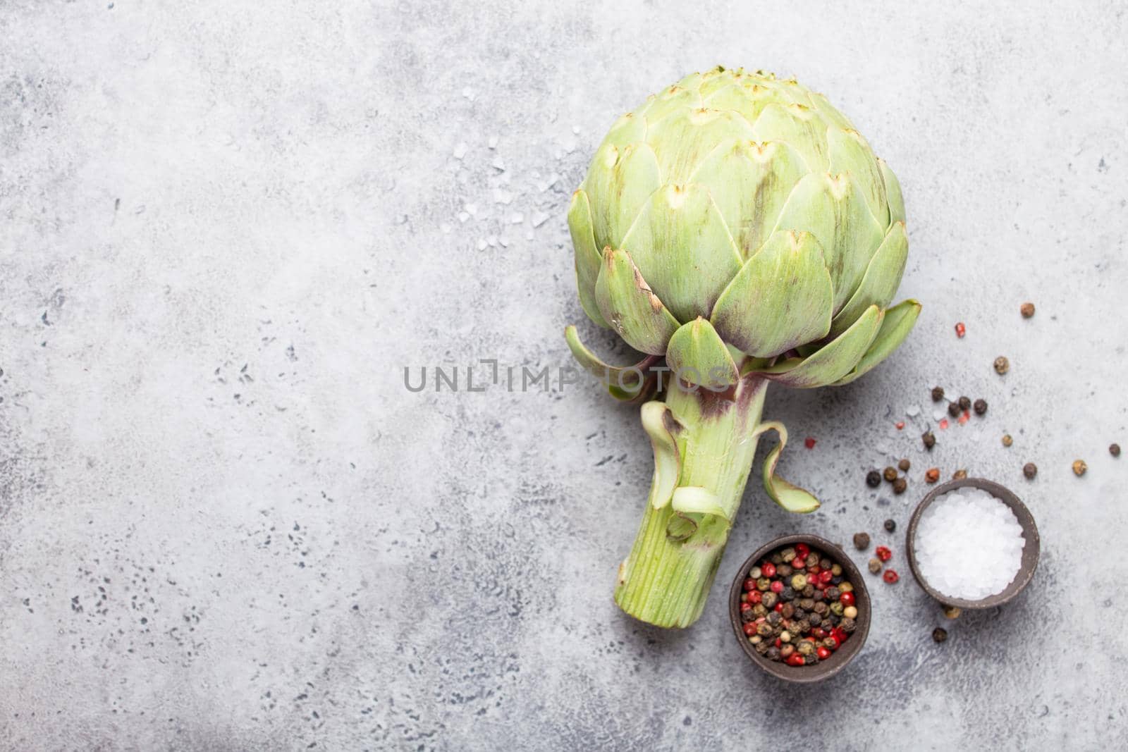 Close-up of raw fresh artichoke with seasonings ready to cook, great as vegetarian food or ingredient for healthy salads and diets, grey stone rustic background, top view. Space for text