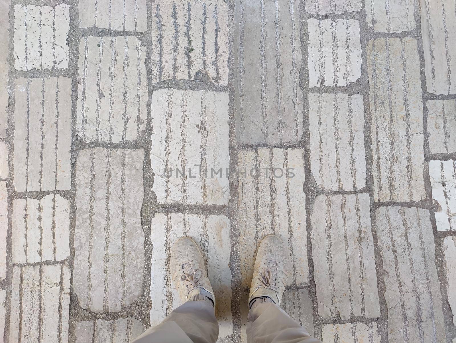 Top down view of male legs in grey trousers and beige sneakers on cobblestone pavement road. Copy space on background