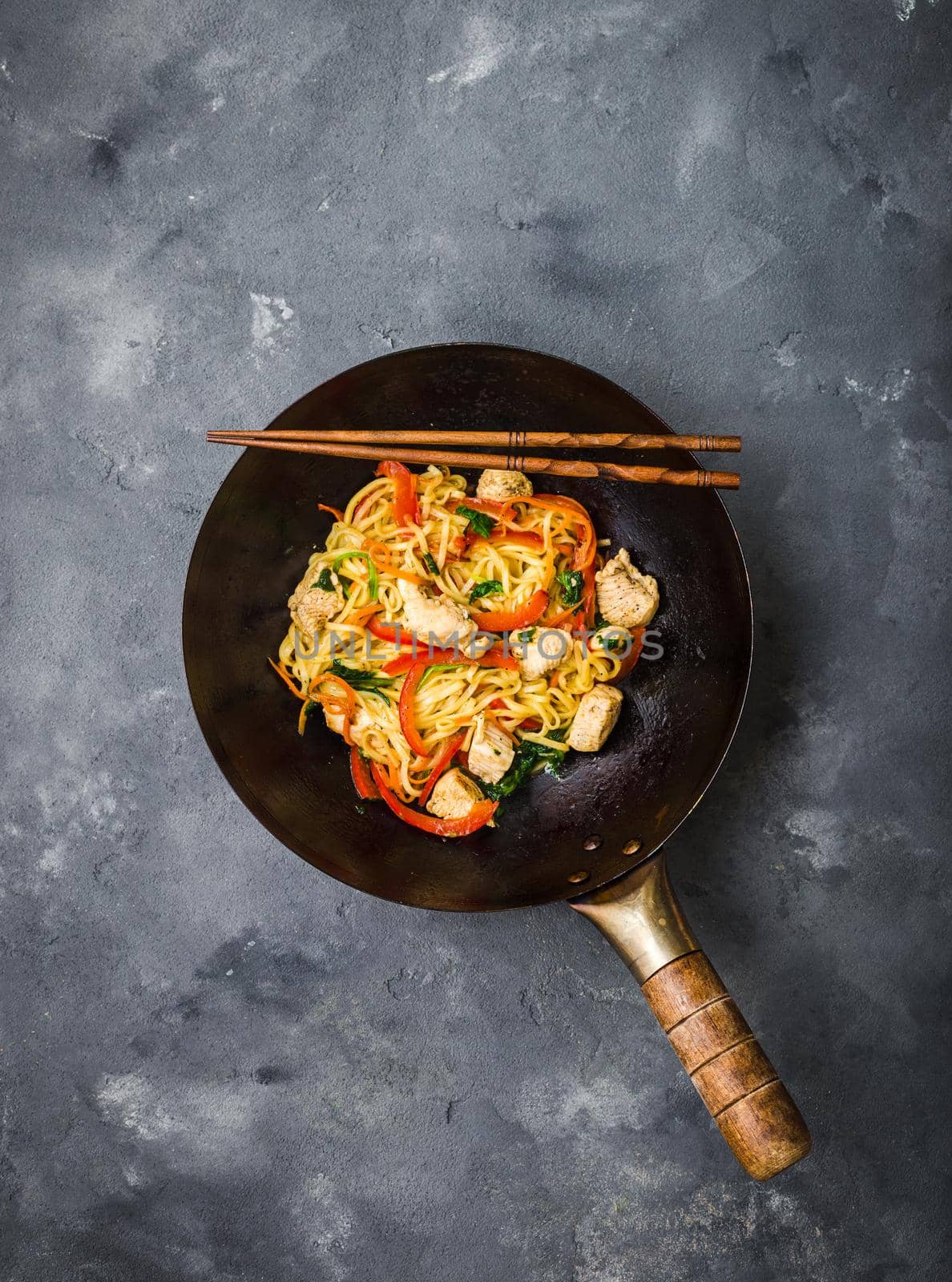 Stir fry noodles, traditional Chinese wok, chopsticks. Asian noodles with vegetables, chicken. Wok noodles, Chinese dinner/lunch. Rustic stone background. Top view. Asian/Chinese noodles. Stir frying.