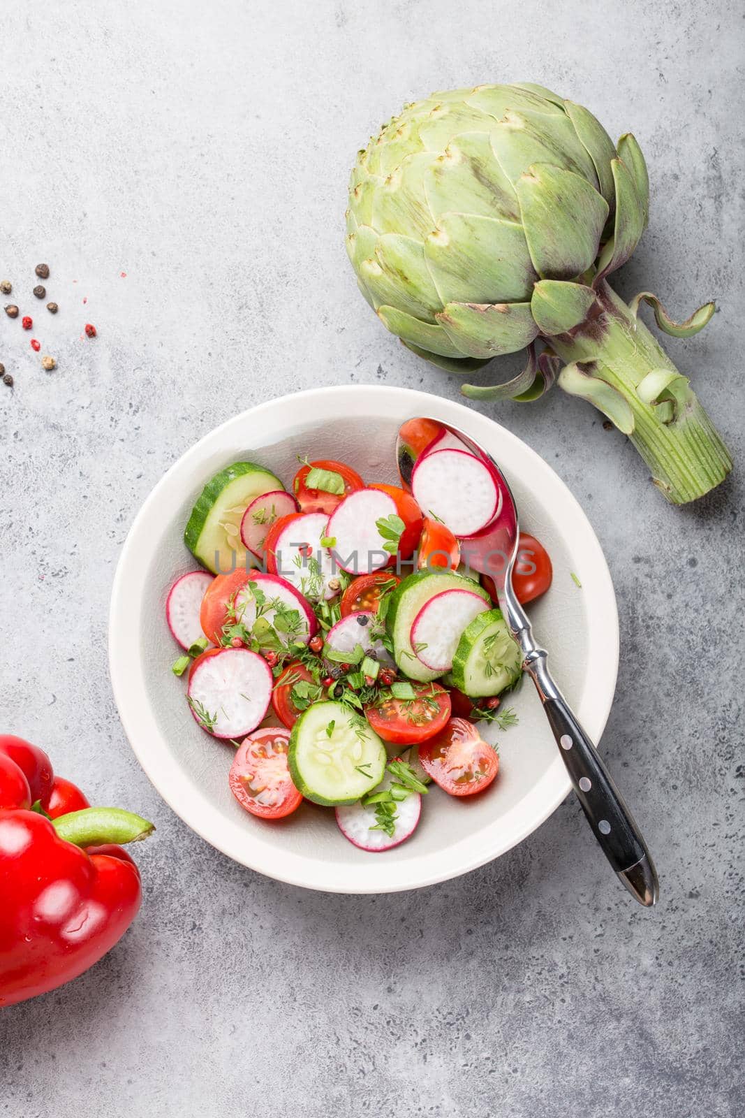 Close-up of fresh healthy salad in a bowl made of tomatoes, cucumber, radish and herbs, with raw artichoke and seasonings, good for diet or detox, grey rustic stone background, top view