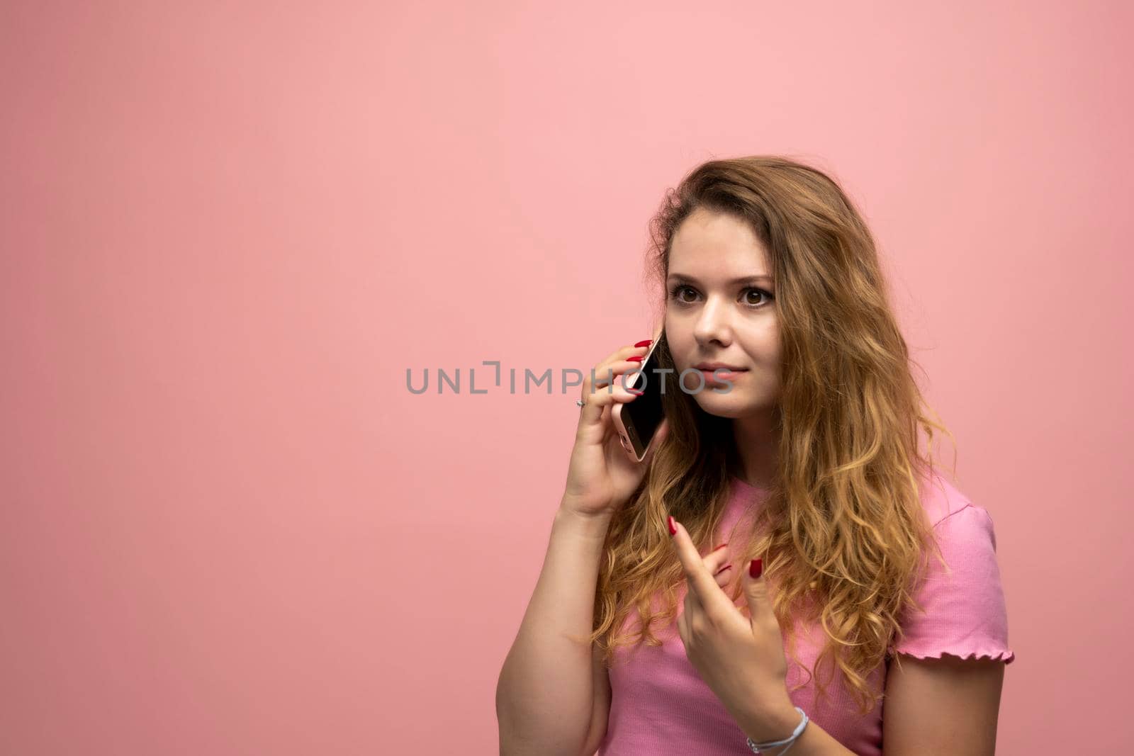 Portrait of a cute happy girl with a curly hair in blue t-shirt talking on mobile phone and laughing isolated over blue background