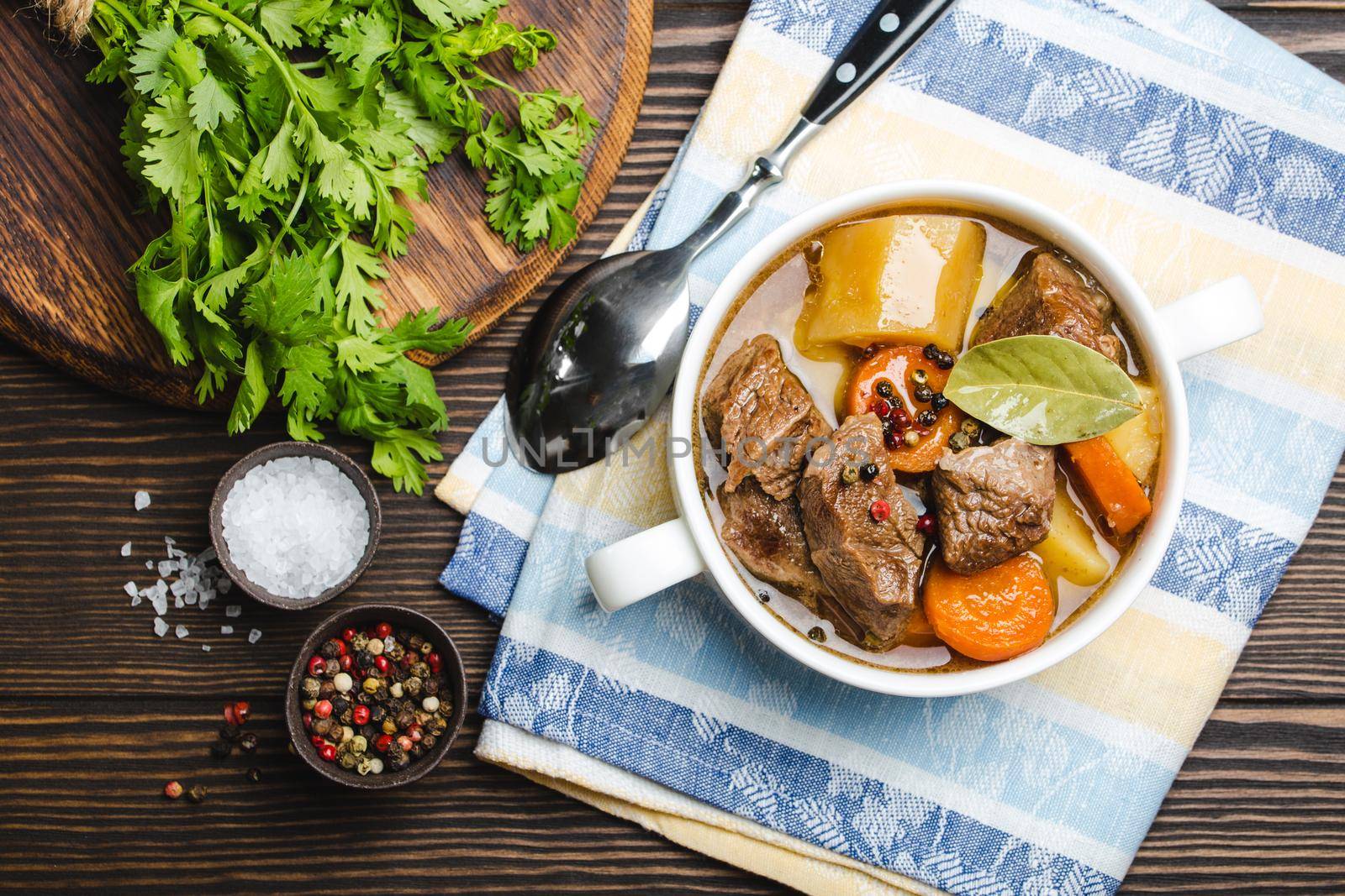 Close-up of slow cooked meat stew ragout in bowl with beef, potato, carrot, broth, herbs, spoon and fork on wooden rustic background, top view. Hot homemade food for dinner, meat casserole .