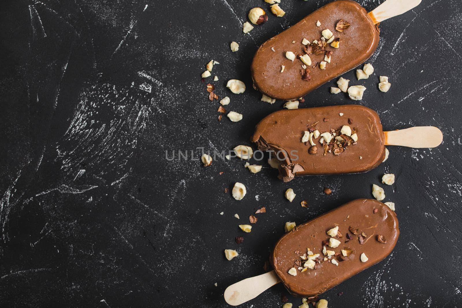 Milk chocolate popsicles with hazelnuts. Close-up. Ice cream popsicles covered with chocolate, sticks, black stone background. Space for text. Top view. Chocolate ice cream bars, nuts. Bitten popsicle