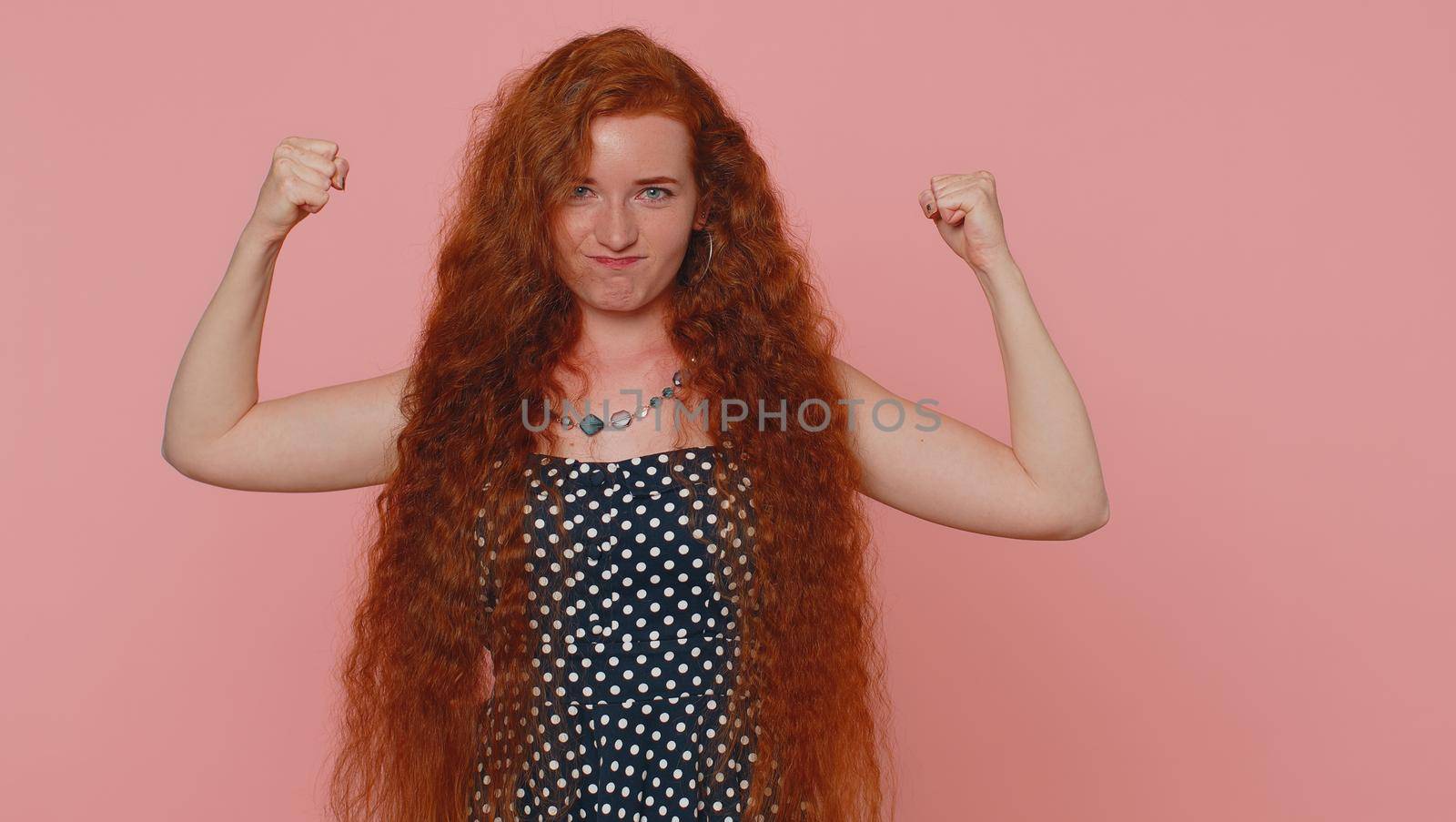 I am strong and independent. Young redhead woman showing biceps and looking confident, feeling power strength to fight for rights, energy to gain success win. Ginger girl on pink studio background