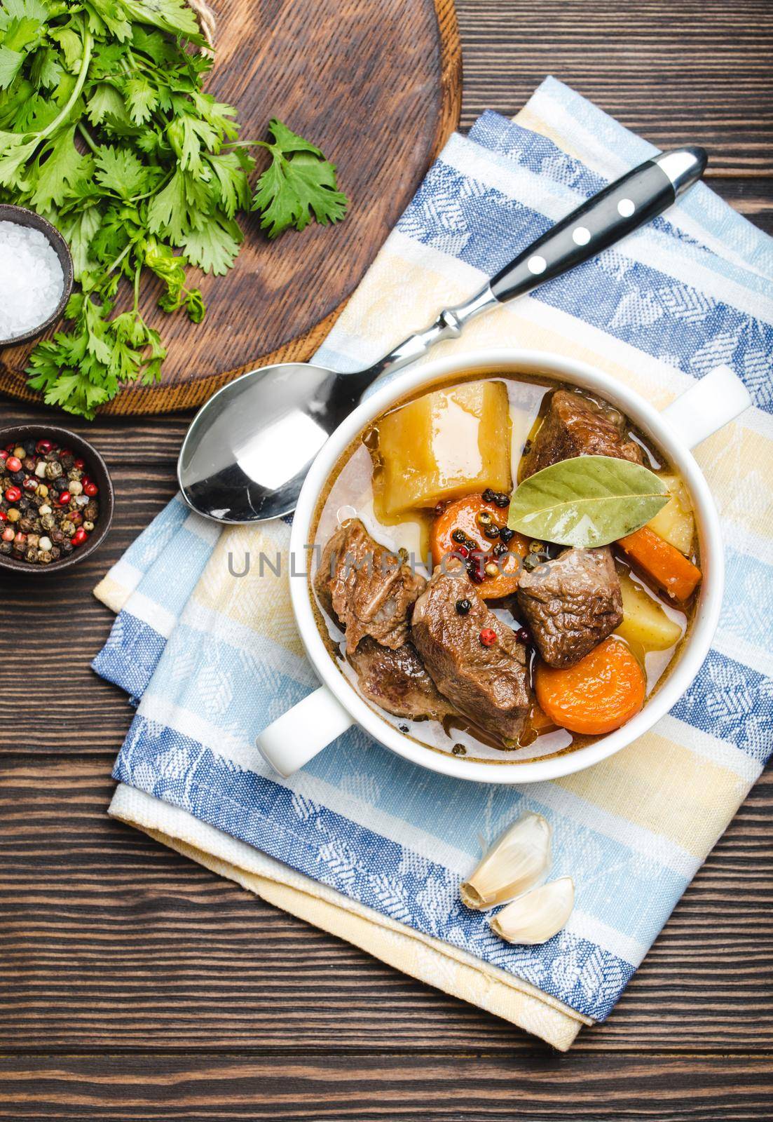 Close-up of slow cooked meat stew ragout in bowl with beef, potato, carrot, broth, herbs and spoon on wooden rustic background, top view. Hot homemade food for dinner, meat casserole .