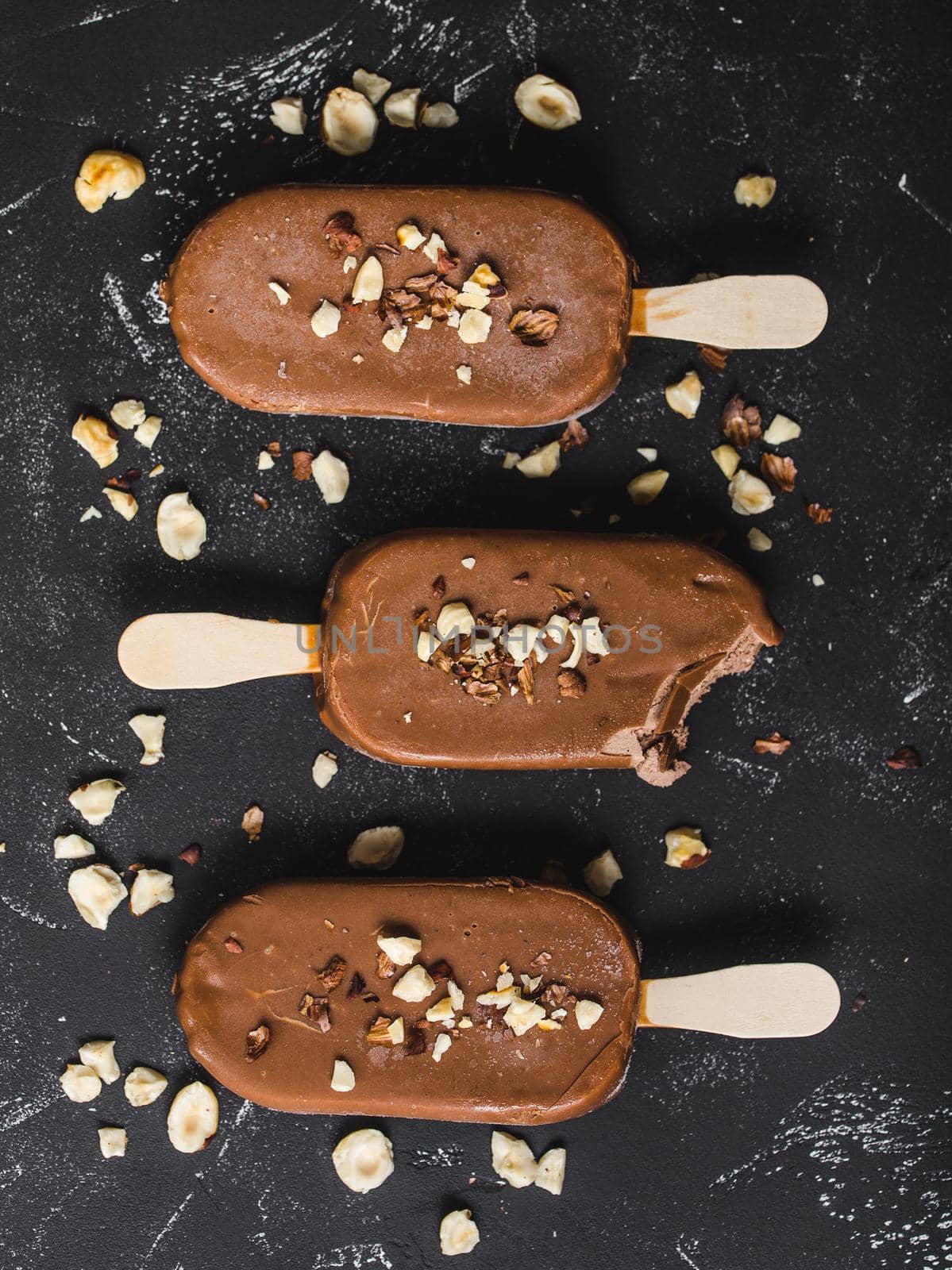 Milk chocolate popsicles with hazelnuts. Close-up. Ice cream popsicles covered with chocolate, sticks, black stone background. Top view. Chocolate ice cream bars, nuts. Dessert. Bitten popsicle