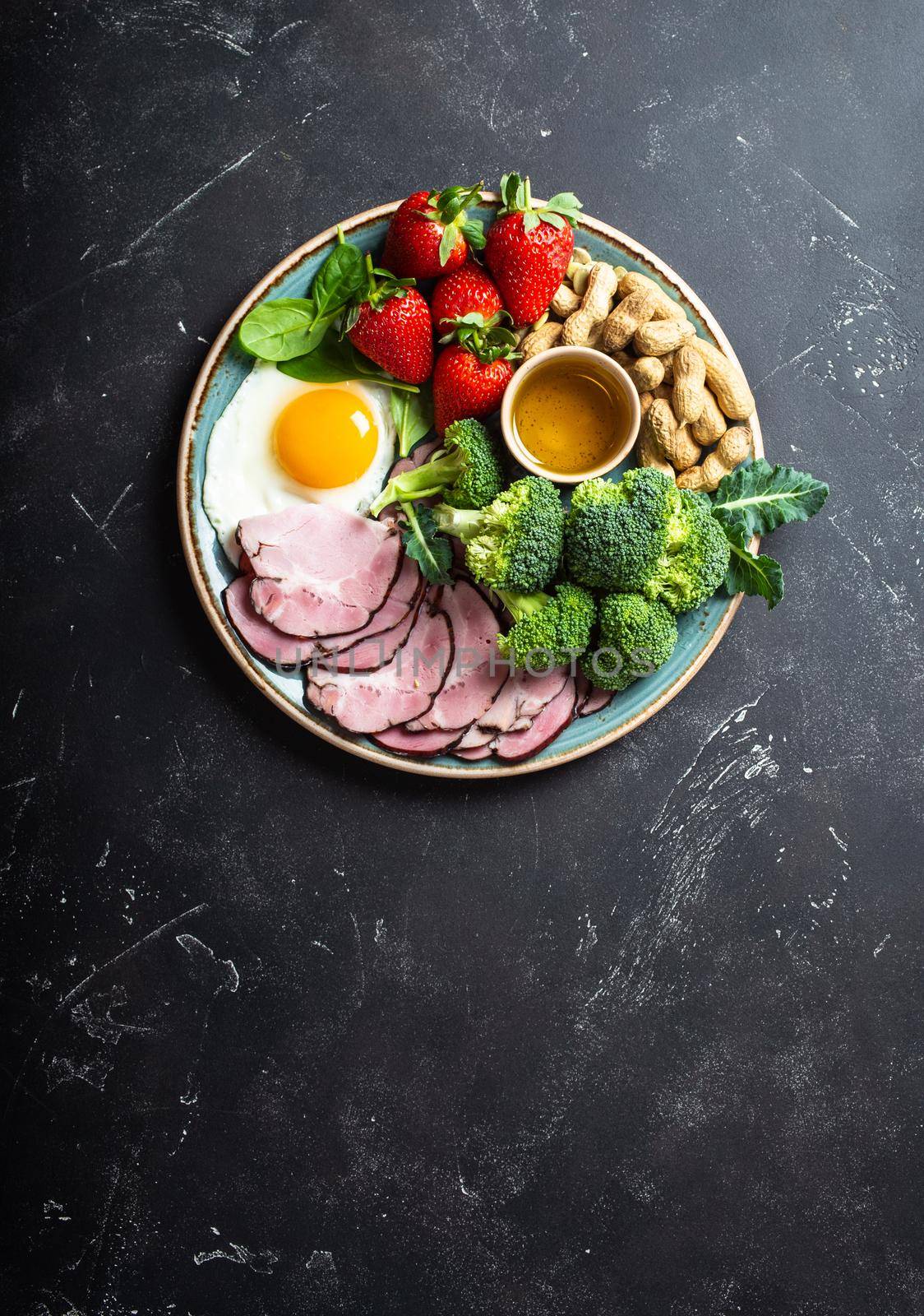 Ketogenic low carbs diet, top view, space for text. Plate on stone black background with keto foods: egg, meat, olive oil, broccoli, berries, nuts, seeds. Healthy fats, clean eating for weight loss