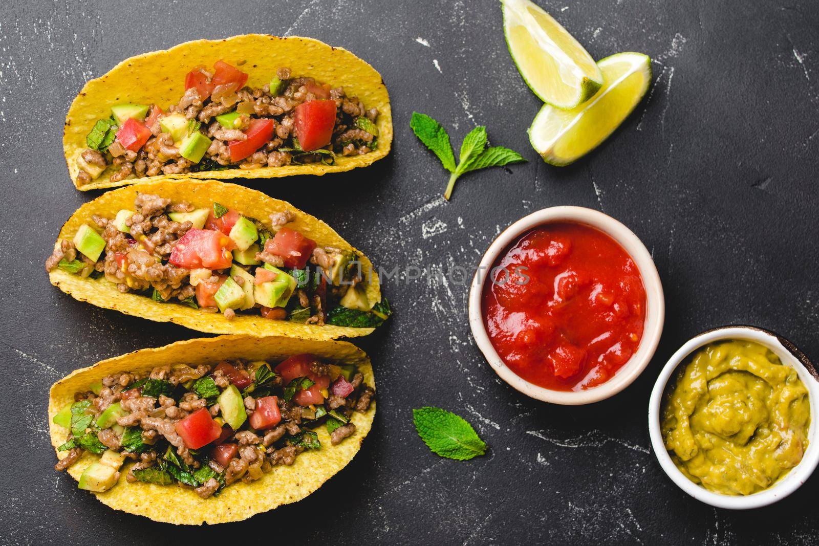 Top view of Mexican tacos with meat, vegetables in yellow corn tortilla served with lime, salsa and guacamole sauces on black rustic stone background. Traditional Mexican dish for lunch or dinner