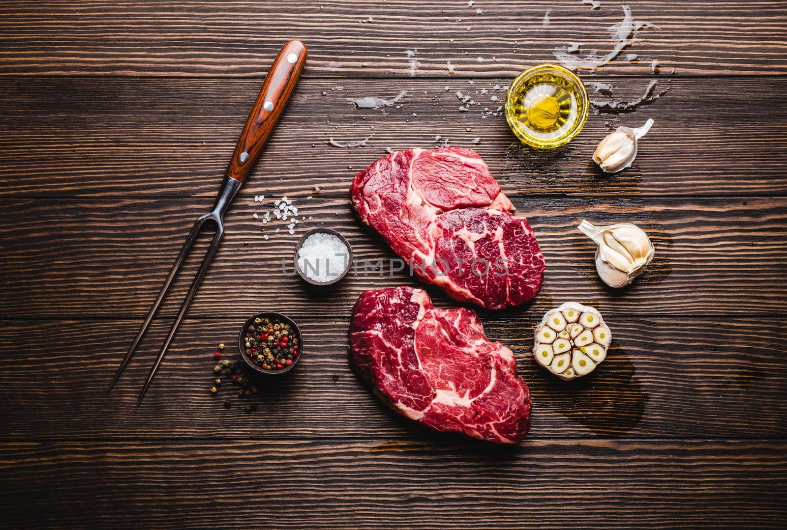 Close-up of raw marbled meat steak Ribeye on rustic wooden background with fork, seasonings and olive oil for cooking, roasting, grill or BBQ party, top view. Meat restaurant concept