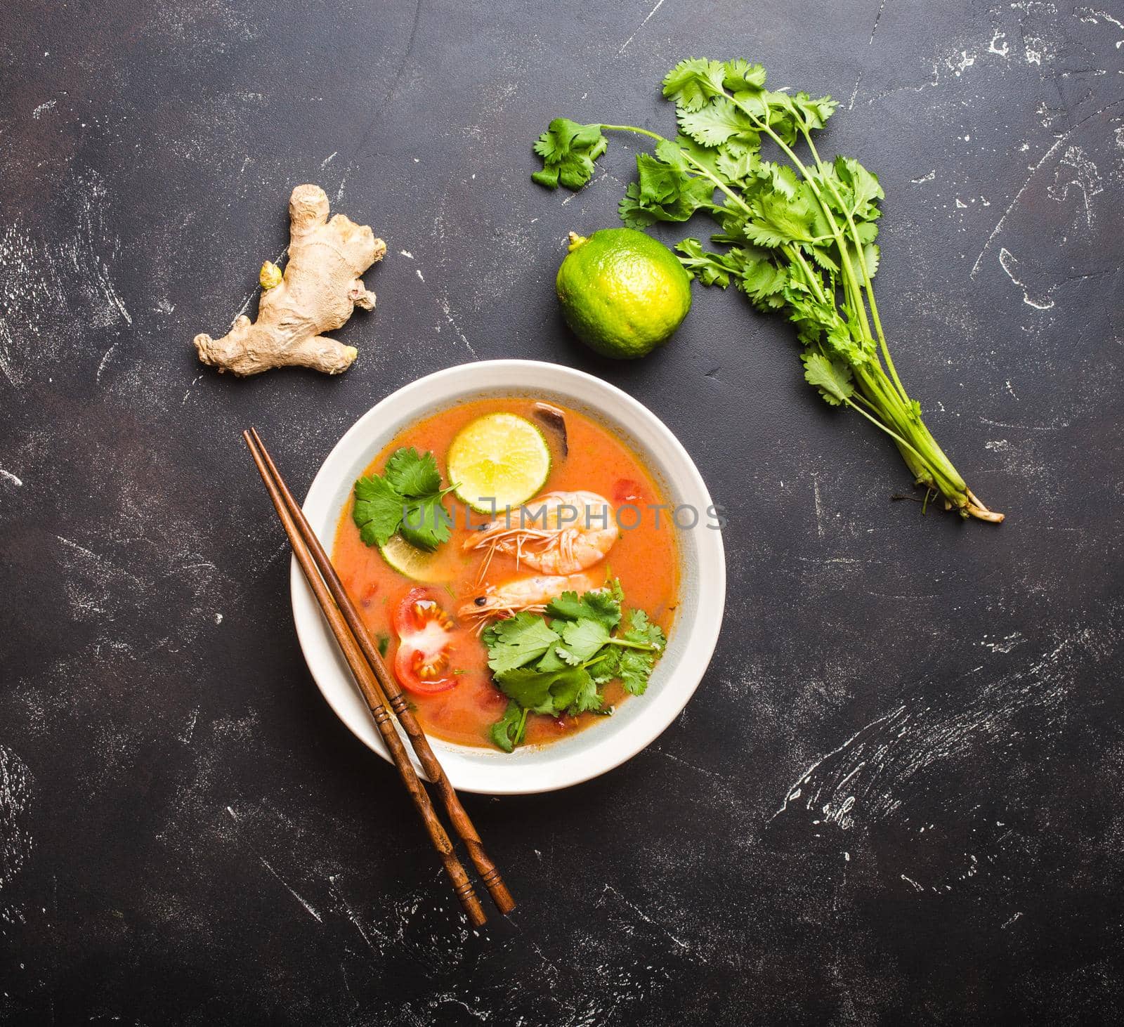 Hot fresh spicy traditional Thai soup tom yum with shrimps, lime, cilantro in a bowl on rustic black stone background, top view. Authentic sweat and sour asian soup for dinner