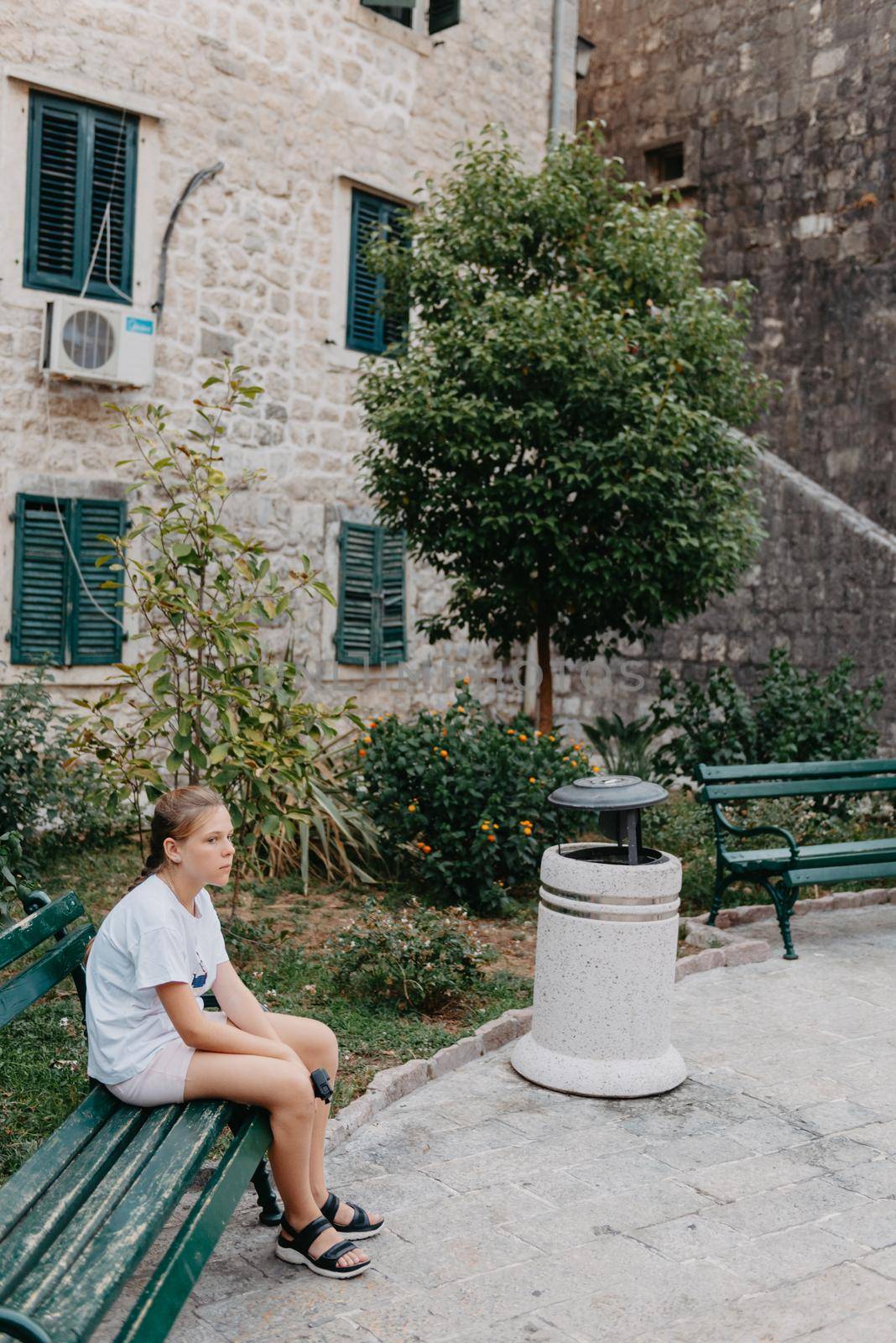 Girl Tourist Resting in the Ancient Narrow Street On A Beautiful Summer Day In MEDITERRANEAN MEDIEVAL CITY, OLD TOWN KOTOR, MONTENEGRO. Young Beautiful Cheerful Woman Walking On Old Street