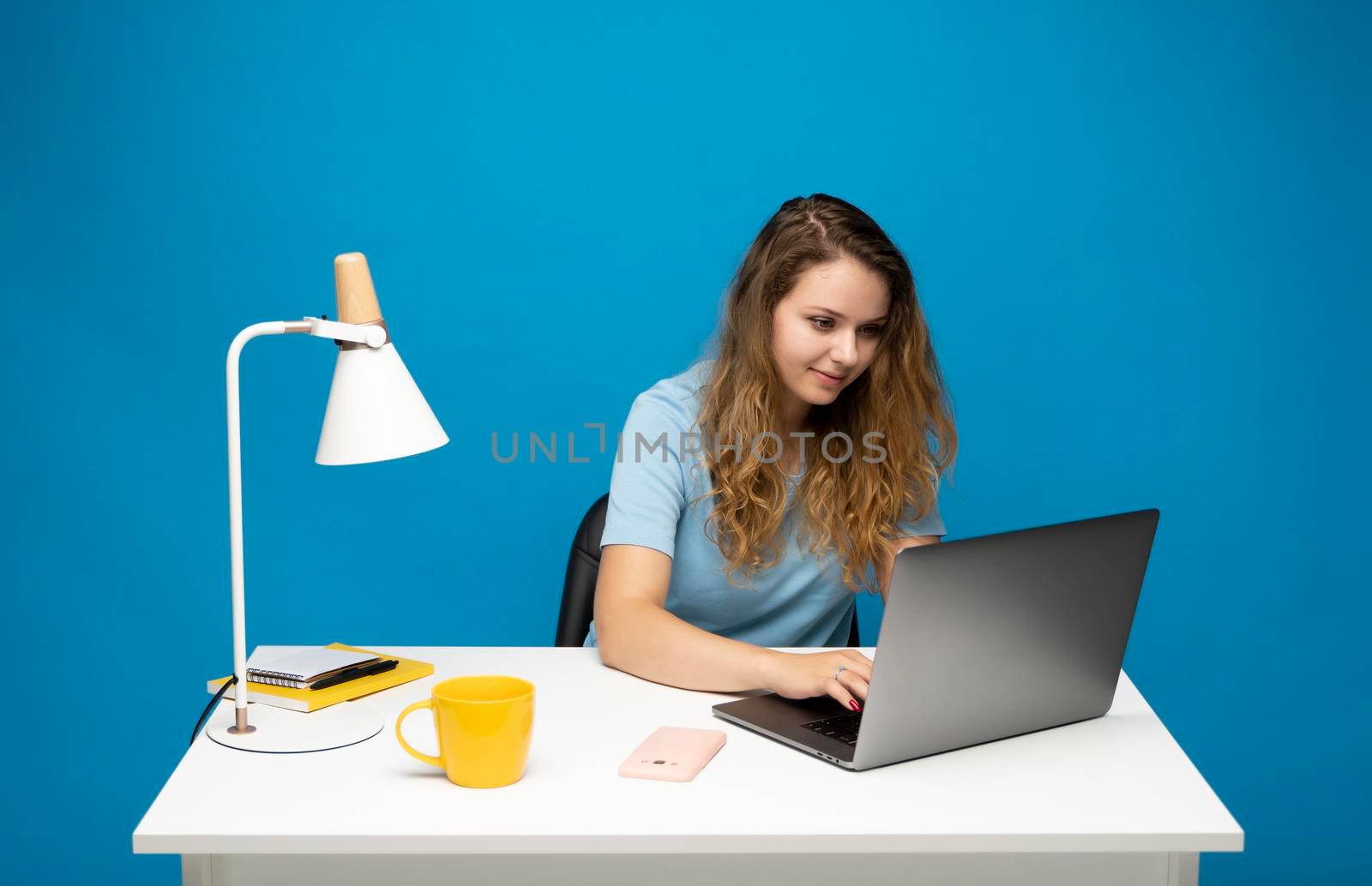 Happy young freelancer curly woman in a blue t-shirt working with a laptop computer and speaking with a client or friends with messengers. Working on a project. Freelance worker