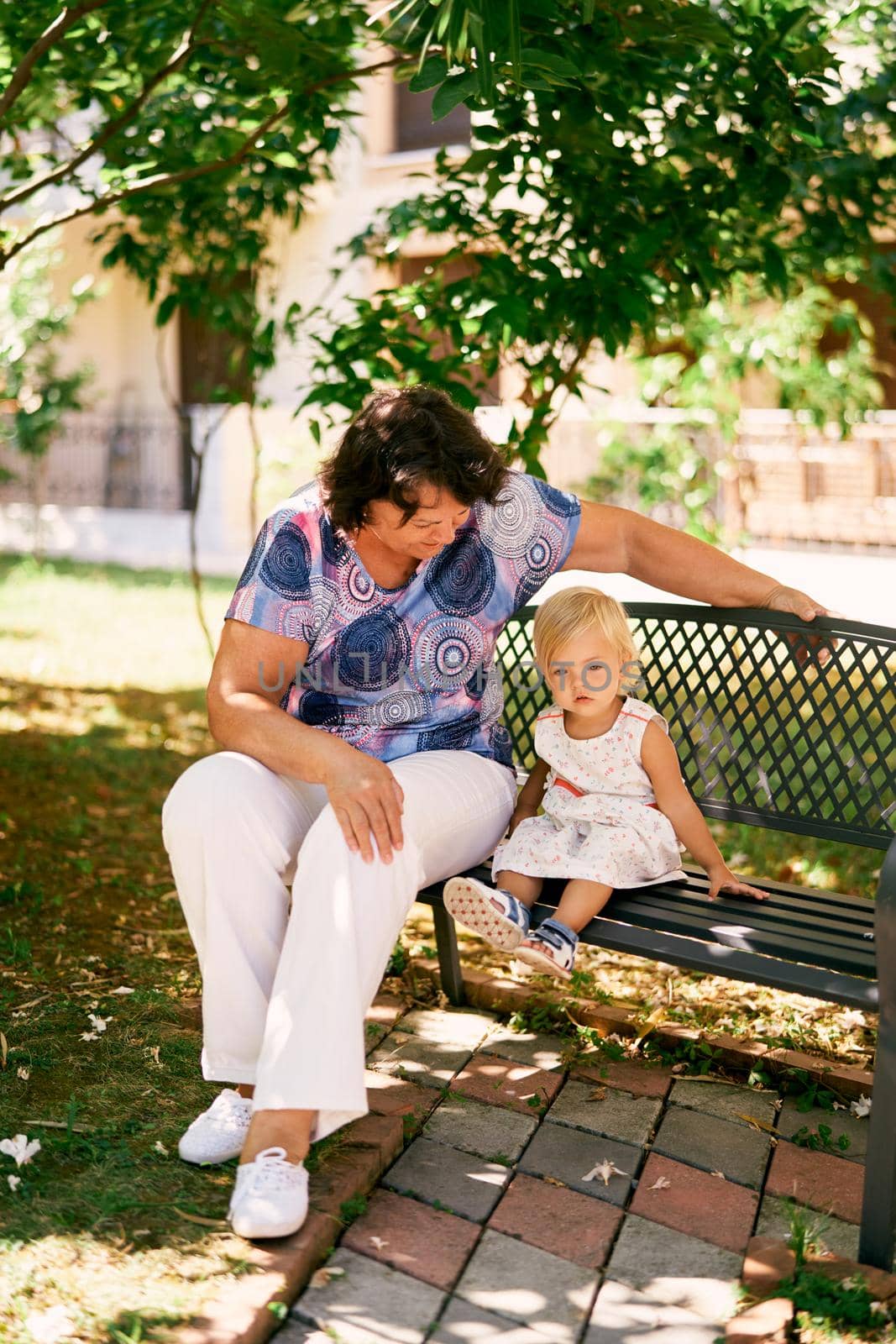 Grandmother sits on a bench next to her little granddaughter by Nadtochiy