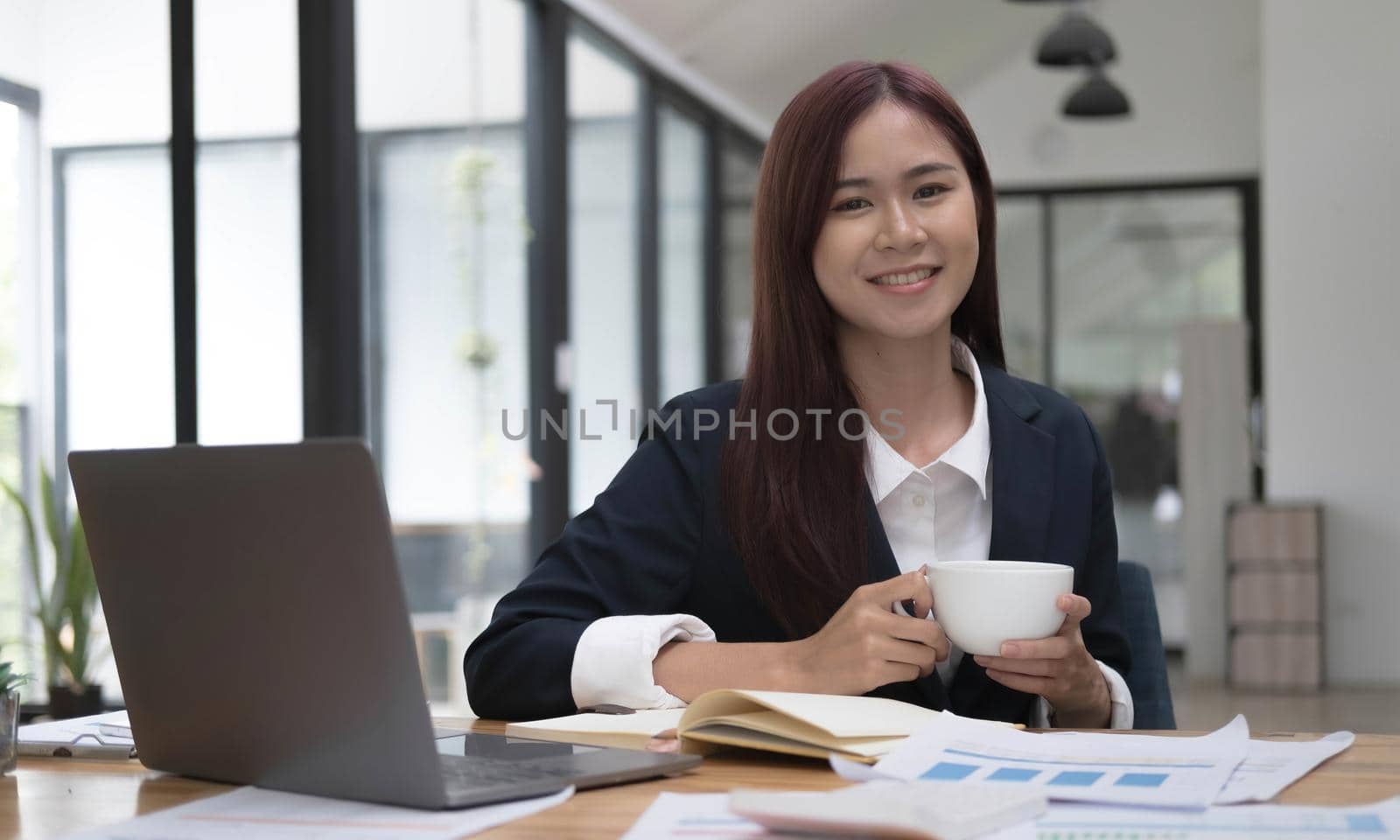 Smiling young Asian businesswoman holding a coffee mug and laptop at the office. Looking at the camera..