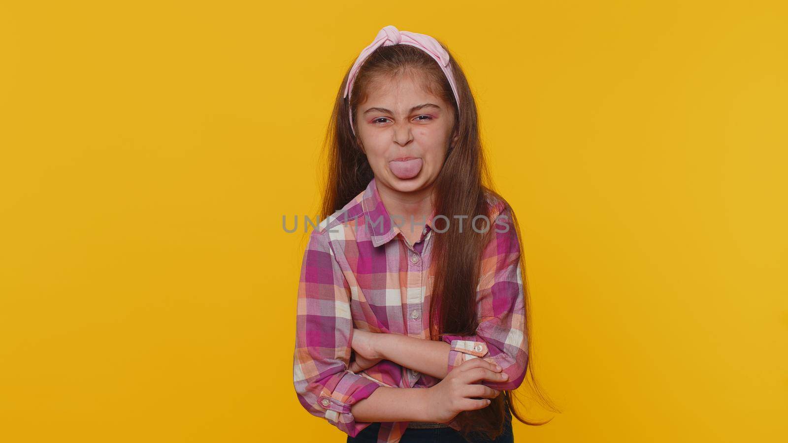 Cheerful funny young preteen child girl kid showing tongue making faces at camera, fooling around, joking, aping with silly face, teasing. Little toddler children isolated on studio yellow background