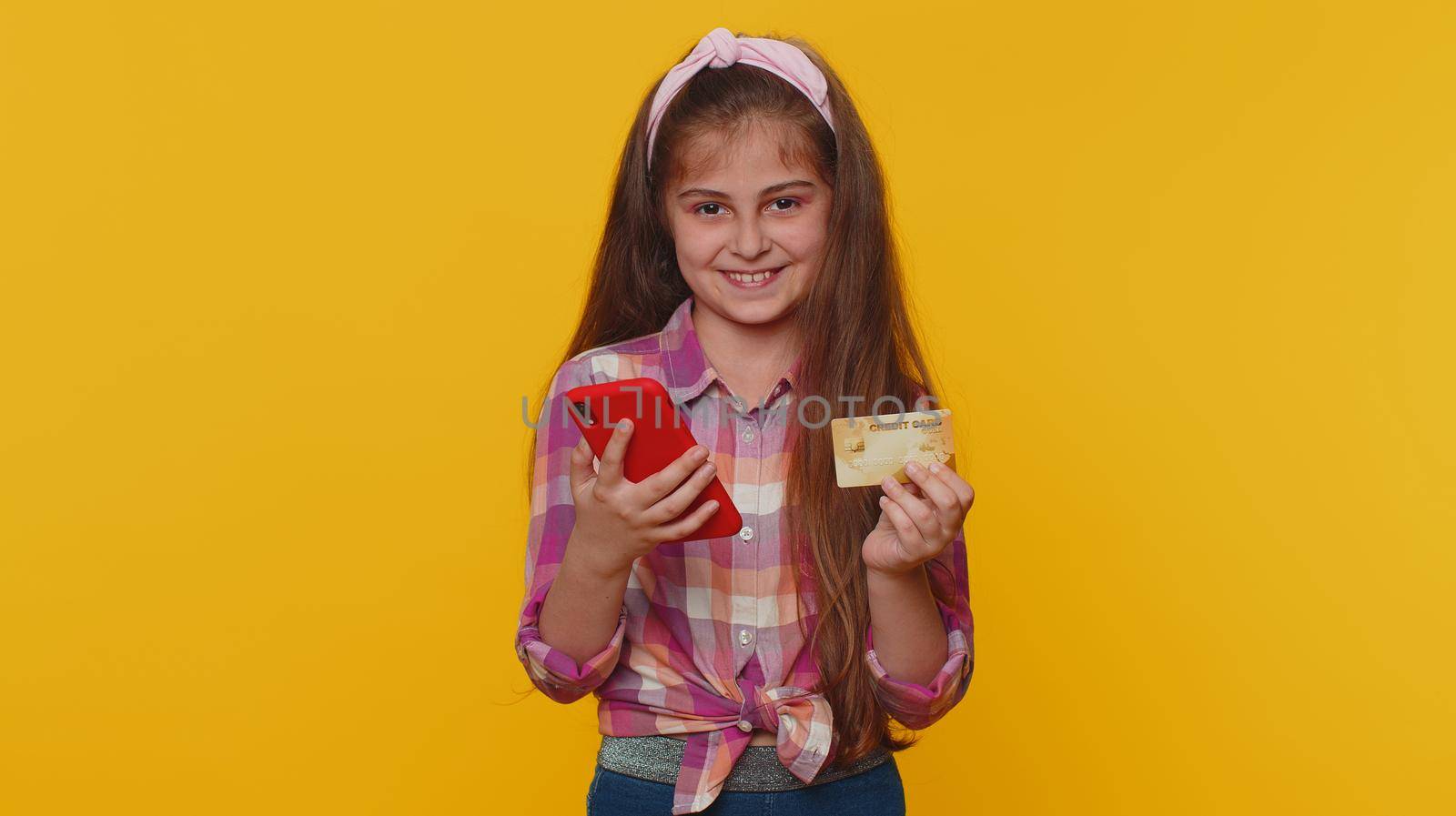 Young preteen child girl kid using credit bank card and smartphone while transferring money, purchases online shopping, order food delivery. Little toddler children alone on studio yellow background