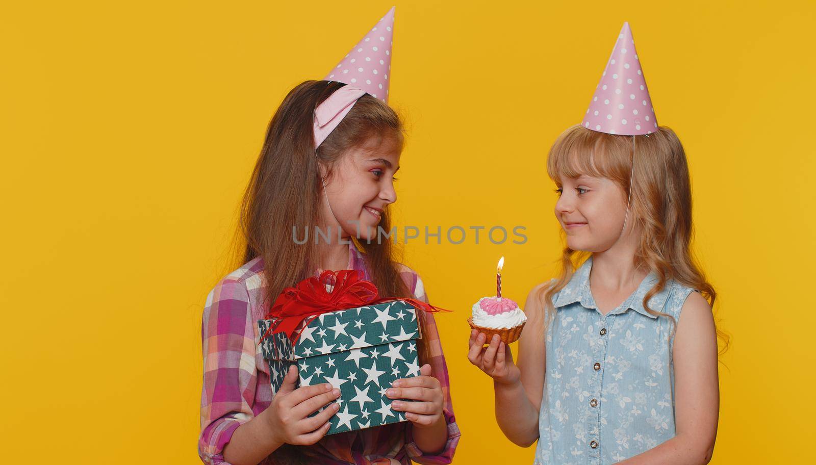 Happy little toddler children sister girls siblings friends wearing festive cone cap celebrating birthday anniversary party, blowing candle on small cake cupcake making a wish, dancing. Preschool kids