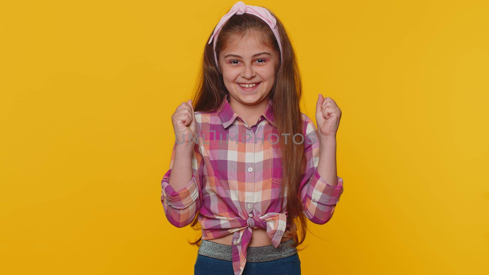 Young preteen child girl kid shouting, raising fists in gesture I did it, celebrating success, winning, birthday, lottery goal achievemen. Little toddler children isolated on studio yellow background