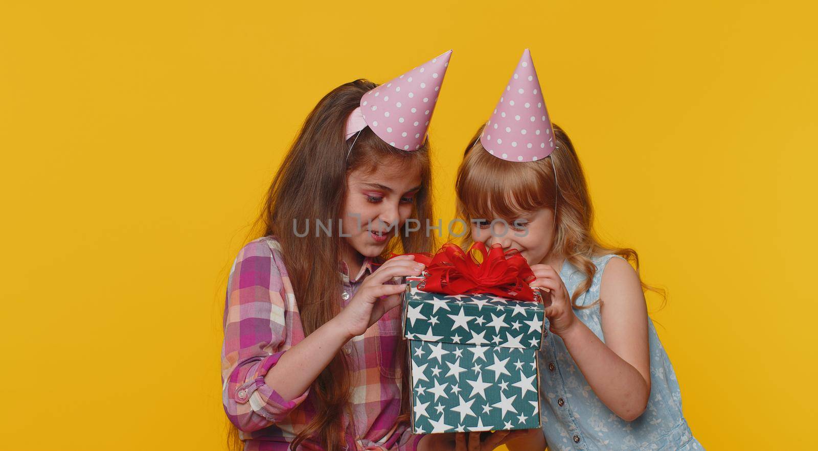 Happy toddler children sisters girls siblings friends wearing festive cone cap celebrating birthday anniversary party, holding gift box. Schoolkids opening present advertising sale discount holidays