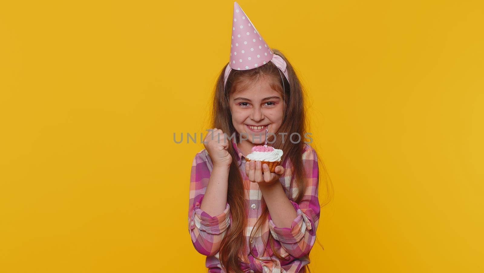 Happy young preteen child girl kid wears festive birthday hat hold cupcake makes wish joyful congratulating blowing burning candle on cake. Little toddler children celebrating anniversary party alone