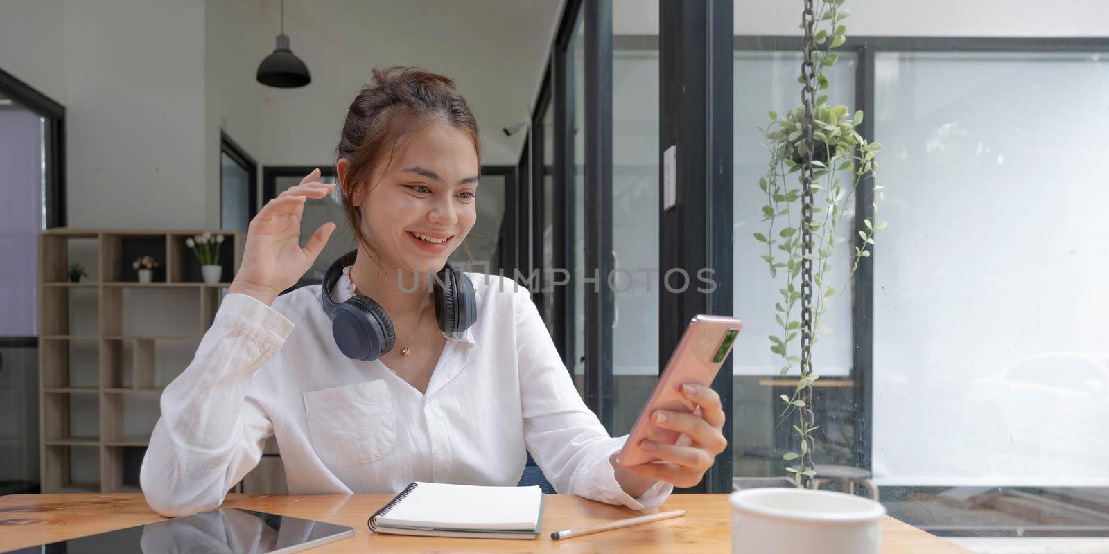 Happy young asian teen girl sit on sofa at home holding phone looking at screen waving hand video calling distance friend online in mobile chat app using smartphone videochat application. by wichayada