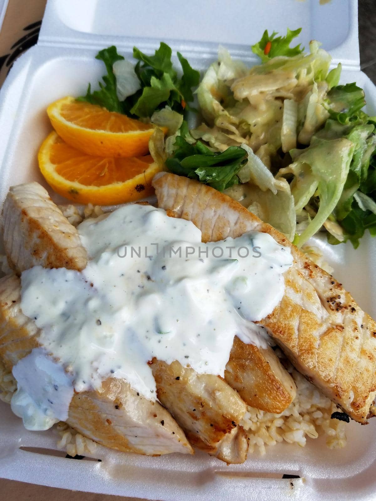 Fish Plate Lunch covered in white sauce with Brown Rice, Orange Slices, and Salad in Styrofoam container.