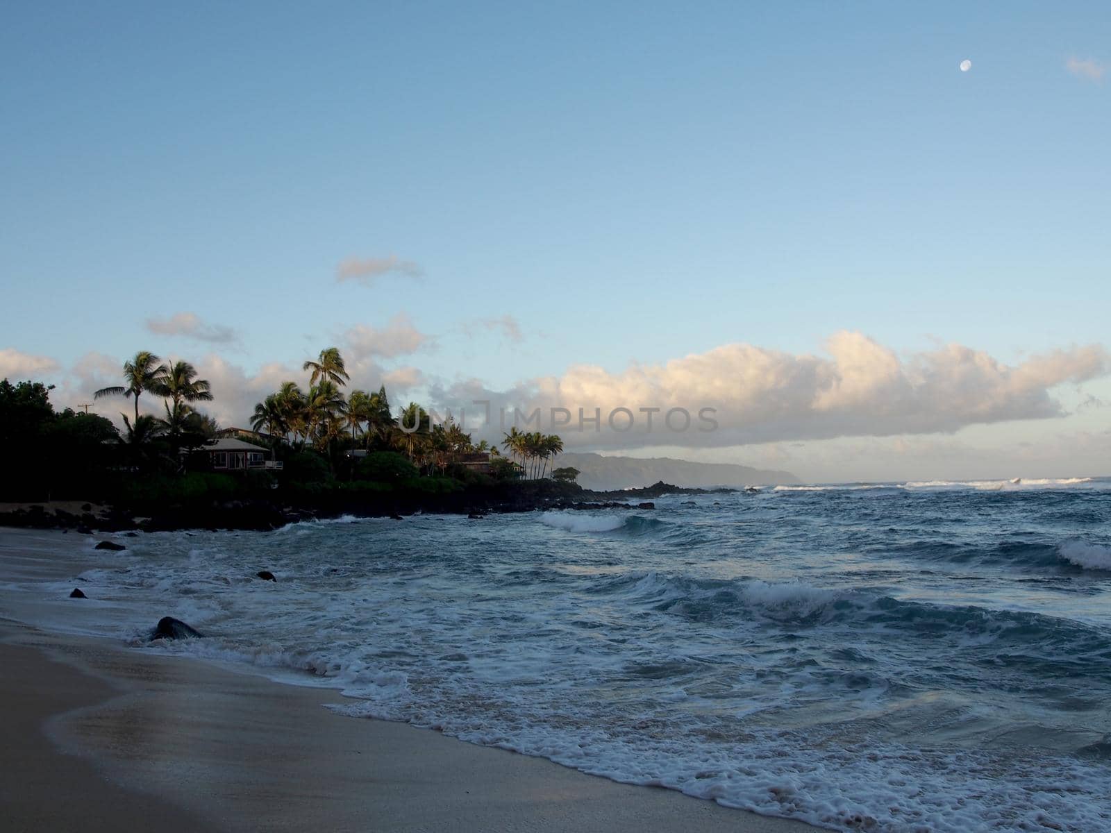 Beautiful Dawn over the ocean with waves crashing into rocks along beach with moon in the sky on the North Shore of Oahu.    