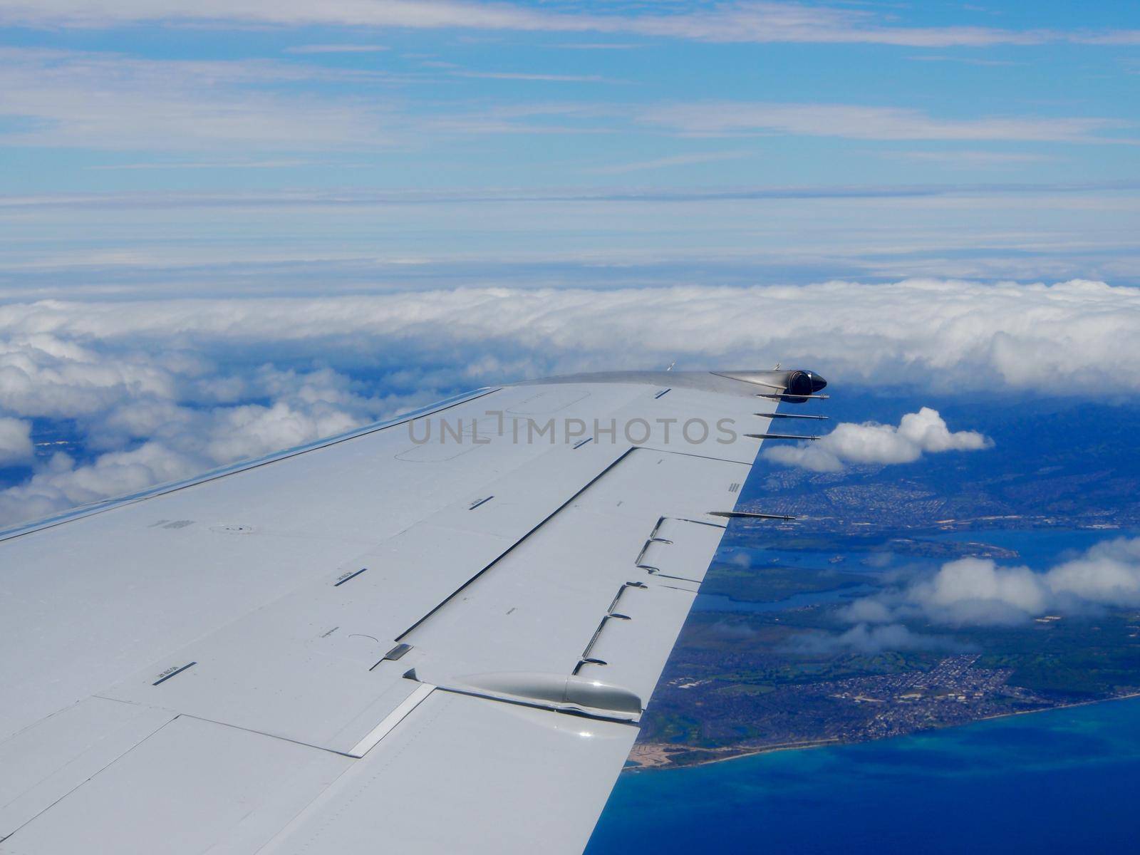 Aerial high in the sky shot of window view of plane leaving Honolulu, Hawaii with the wing of a commercial jet plane with Pearl Harbor, Coast, and clouds of Oahu visible.