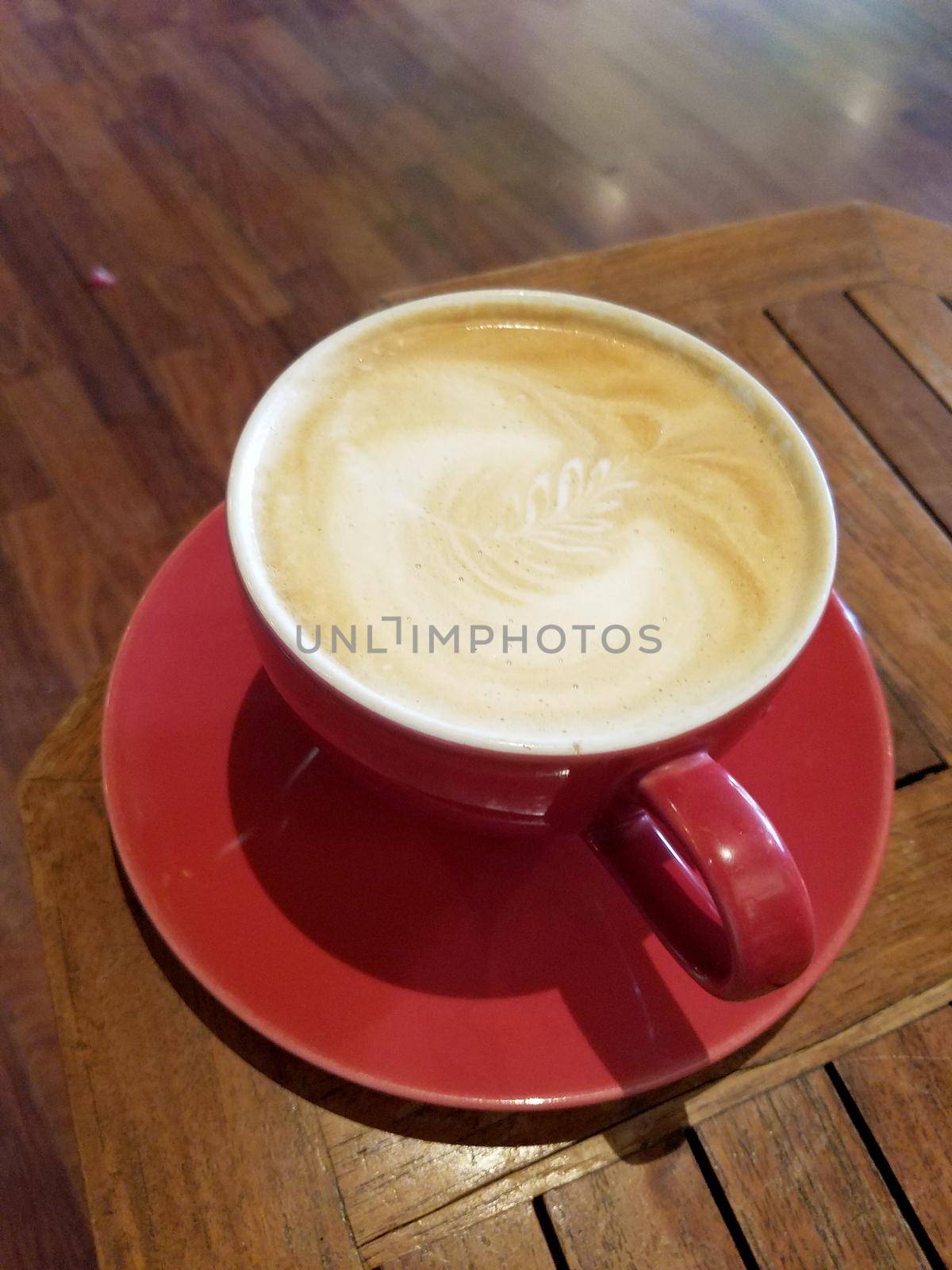 Red cup of Cappuccino on saucer with a leaf pattern in foam on table.