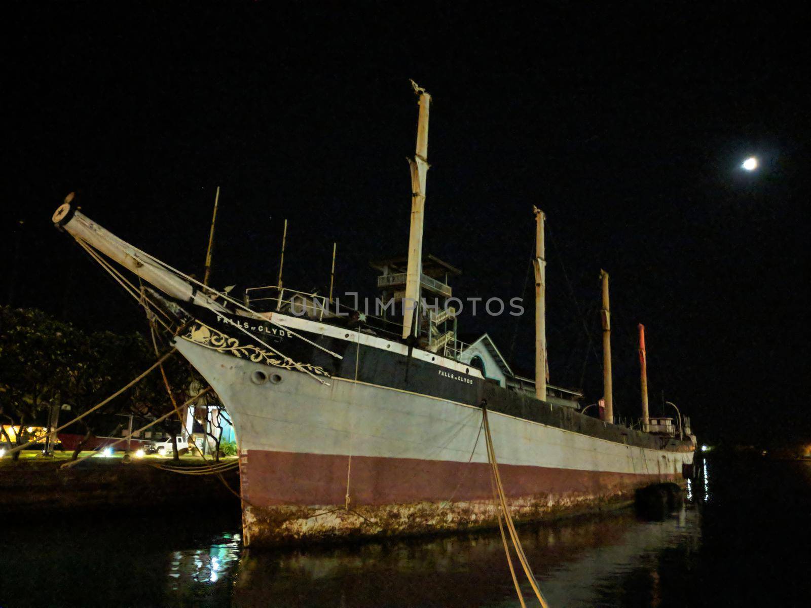 Historic Falls of Clyde Ship sits in Honolulu Harbor at Night with moon in sky. Falls of Clyde is the last surviving iron-hulled, four-masted full rigged ship, and the only remaining sail-driven oil tanker. Designated a U.S. National Historic Landmark in 1989