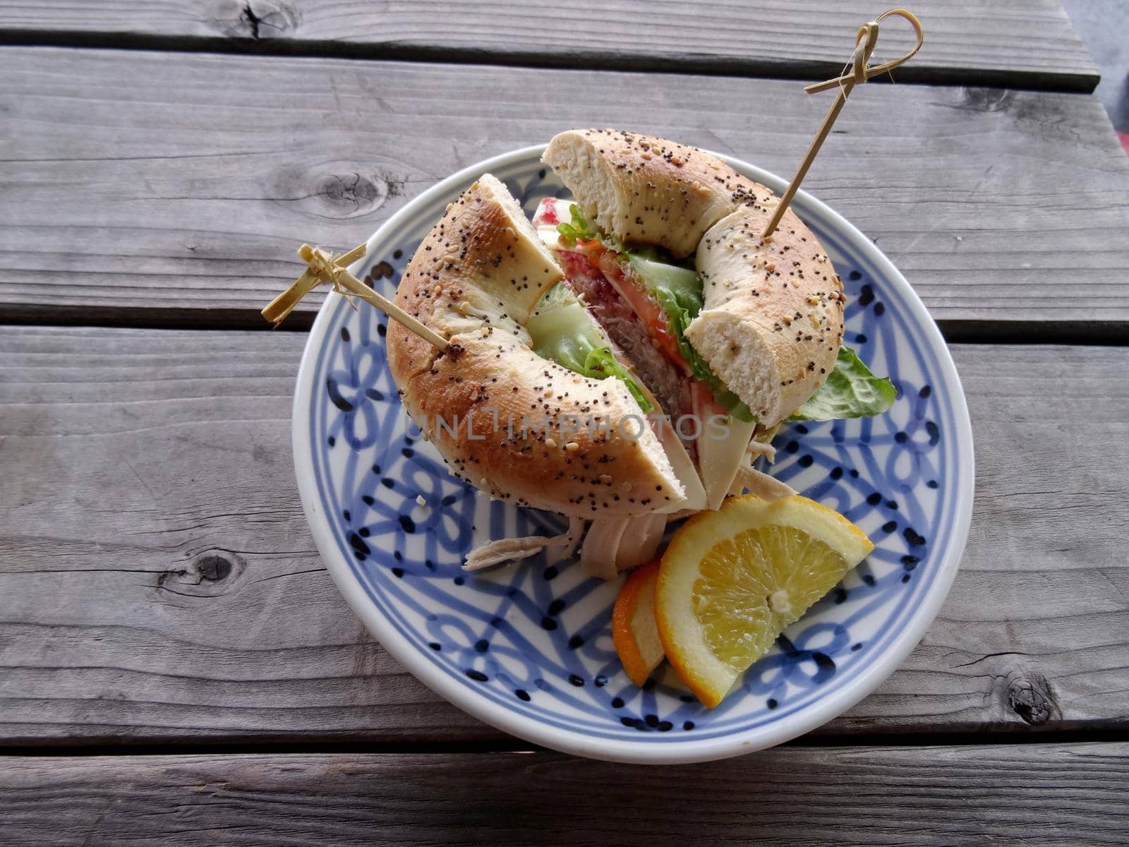 Turkey everything bagel sandwich featuring lettuce, tomato, cheese with orange slices on a plate on a wooden table.