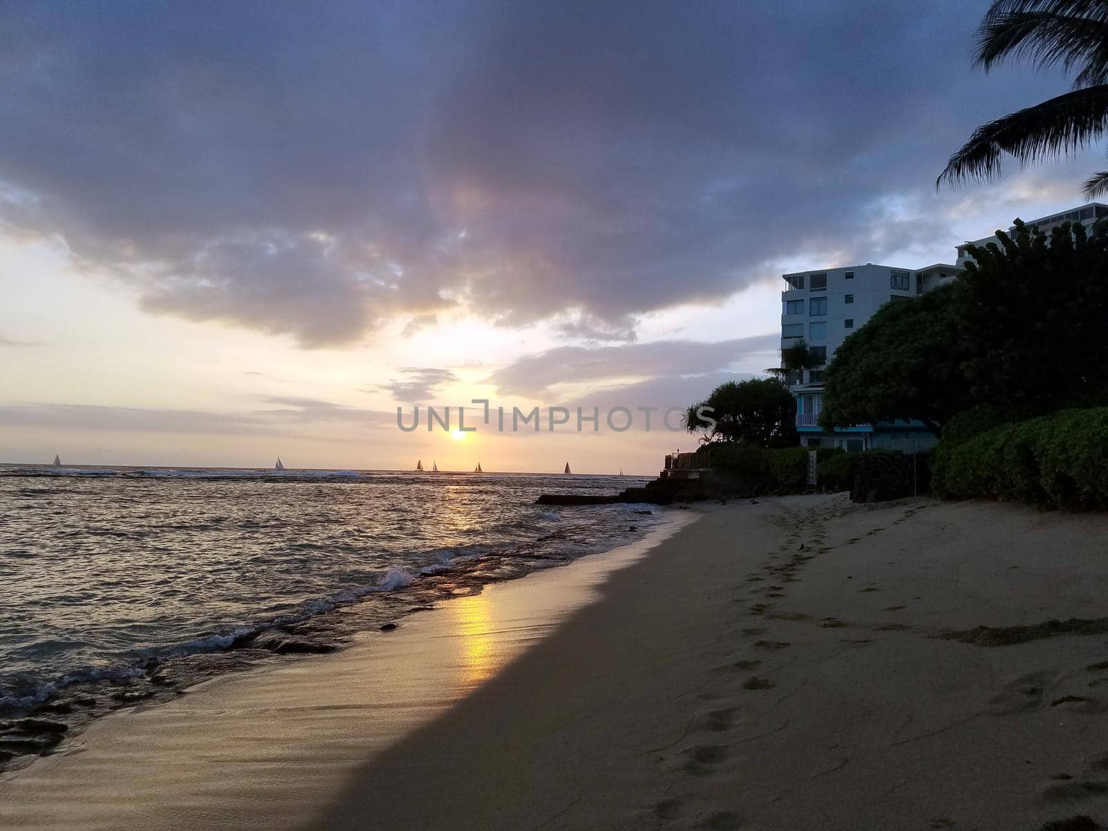Sunsets over Waikiki waters as waves roll into shore at Makalei Beach Park with sail boats in the ocean on Oahu, Hawaii.