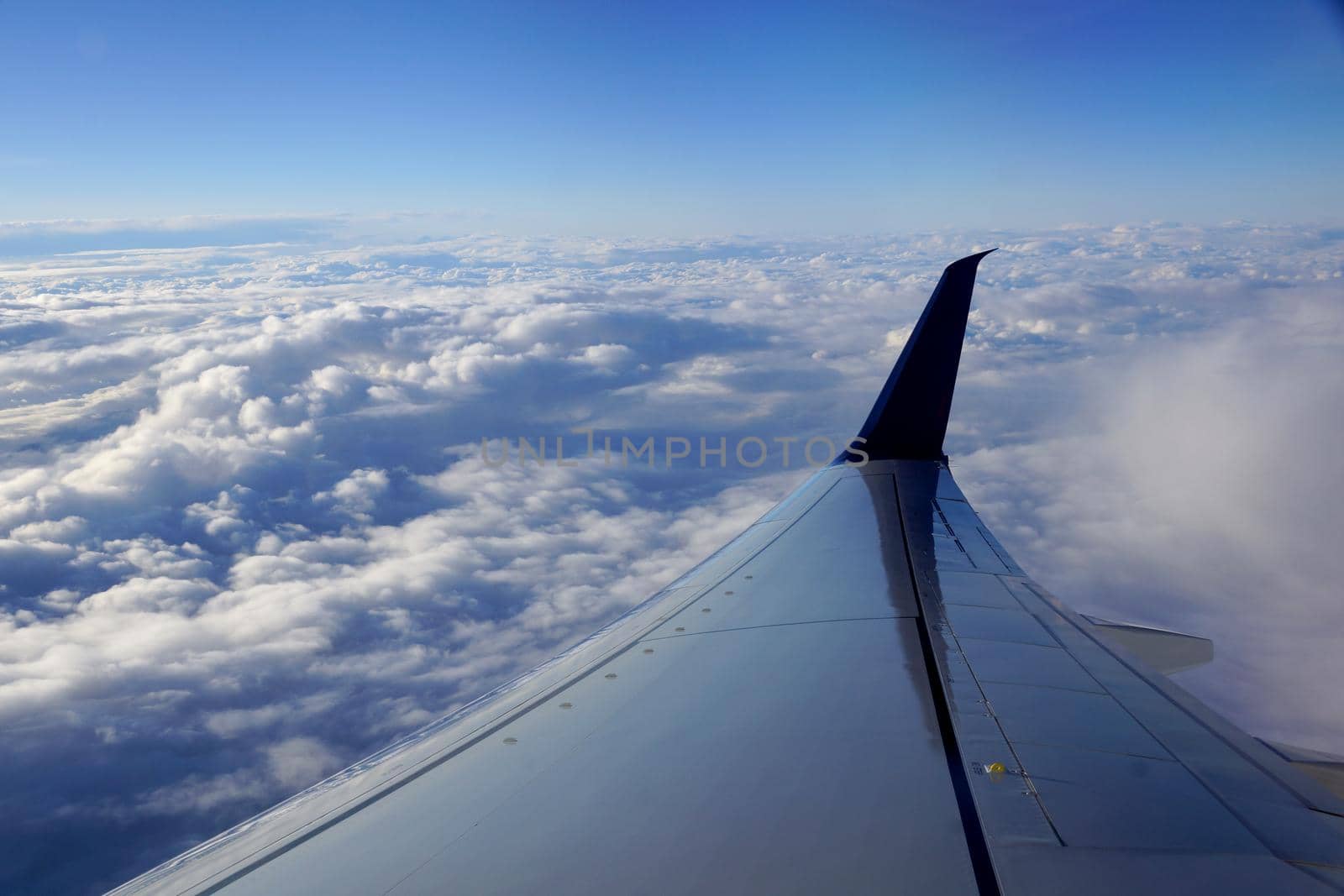 Aerial high in the sky, shot from above the clouds, with the wing of a commercial jet plane that turns upwards on the right side and blue skys.