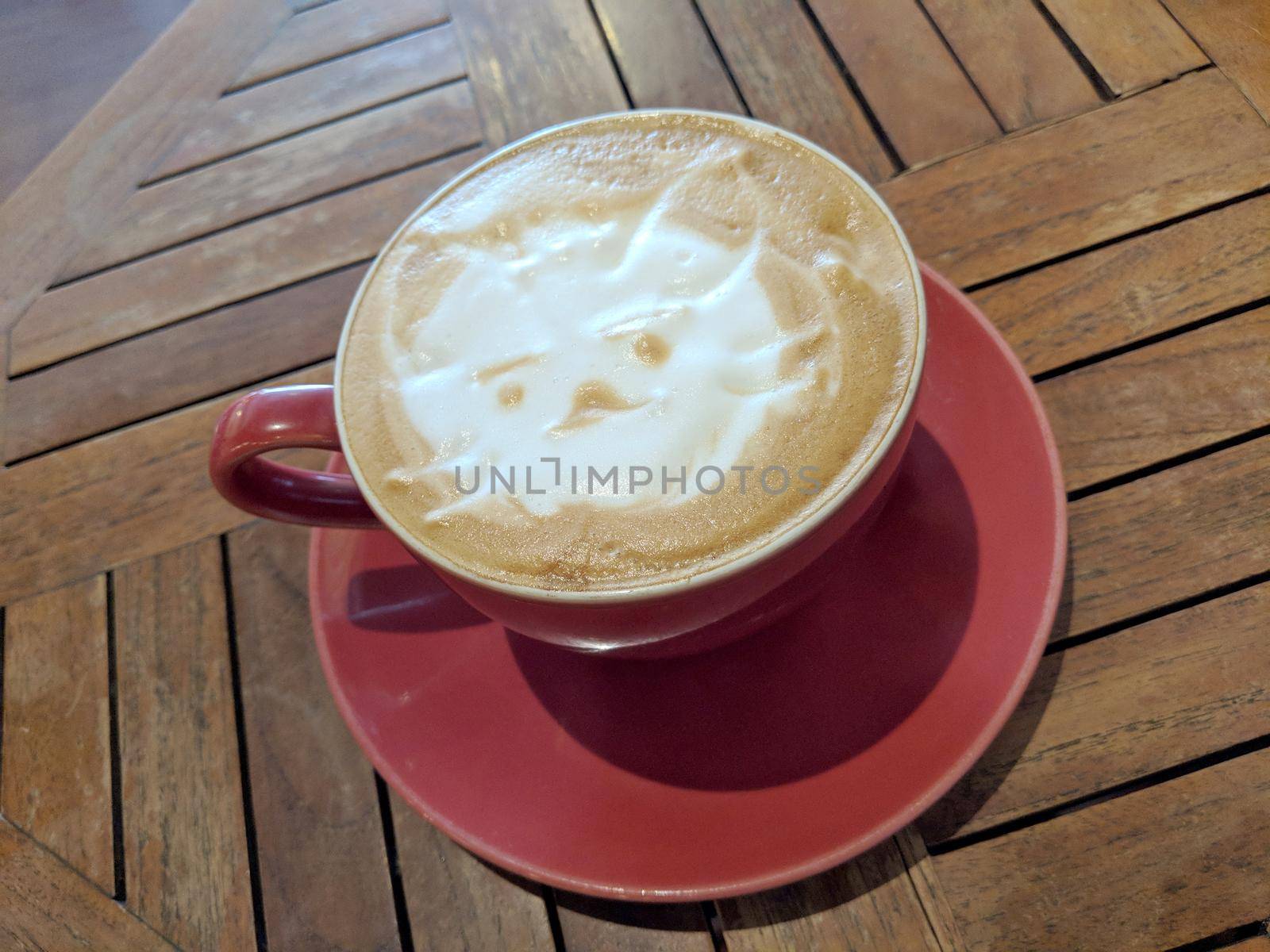 Red cup of Cappuccino on saucer with a cat face pattern in foam on table.